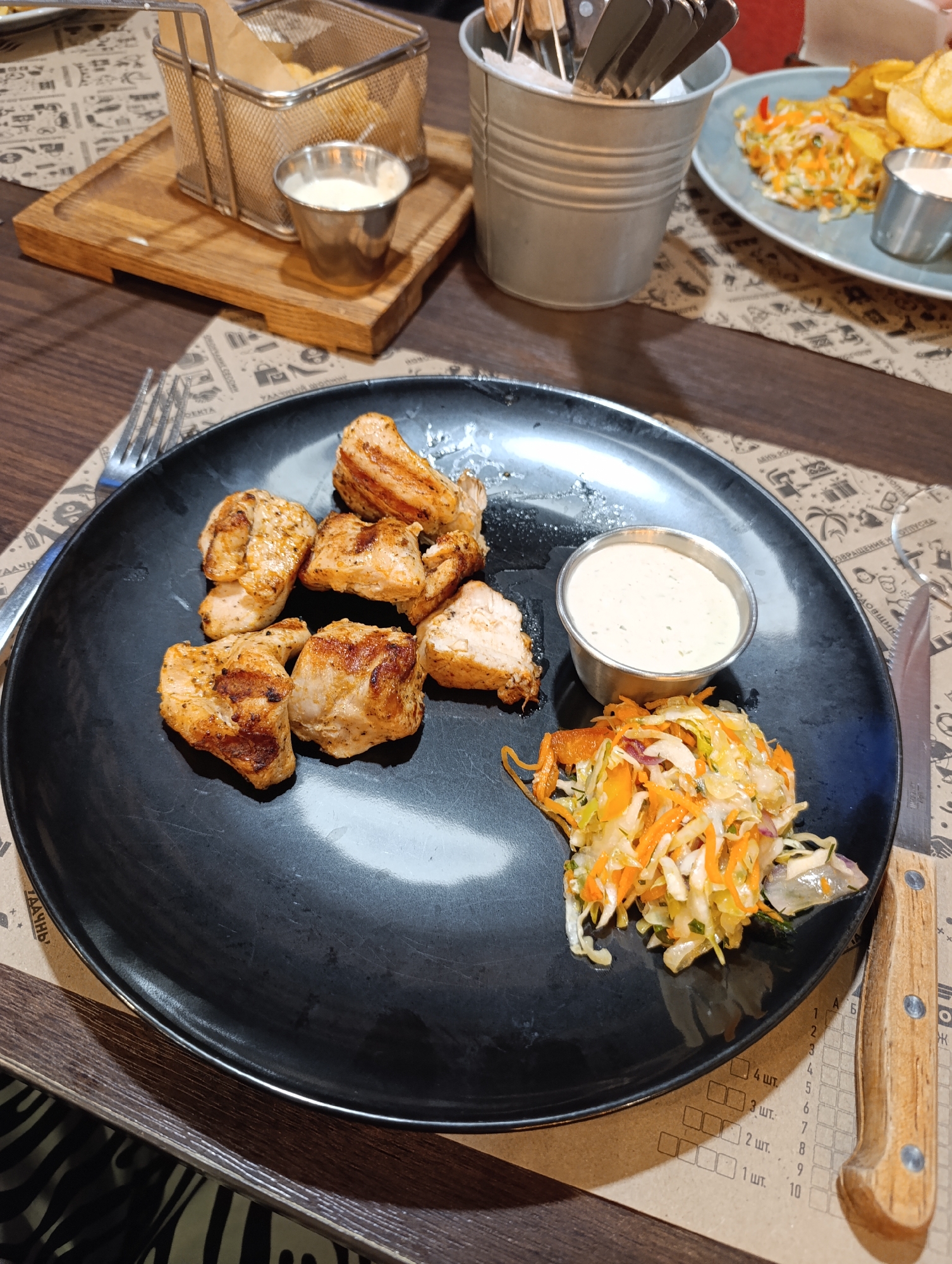The image depicts a plate of food on a table. The plate is black and contains pieces of grilled meat, a small bowl of white sauce, and a small portion of coleslaw. To the left of the plate is a fork, and to the right is a knife. In the background, there's a wire basket containing what appears to be fried potatoes, a small bowl of white sauce next to it, a metal container holding utensils, and another plate with food on it. The table has a printed placemat underneath the plates. The location in the city of Unknown, Unknown is not ascertainable.
