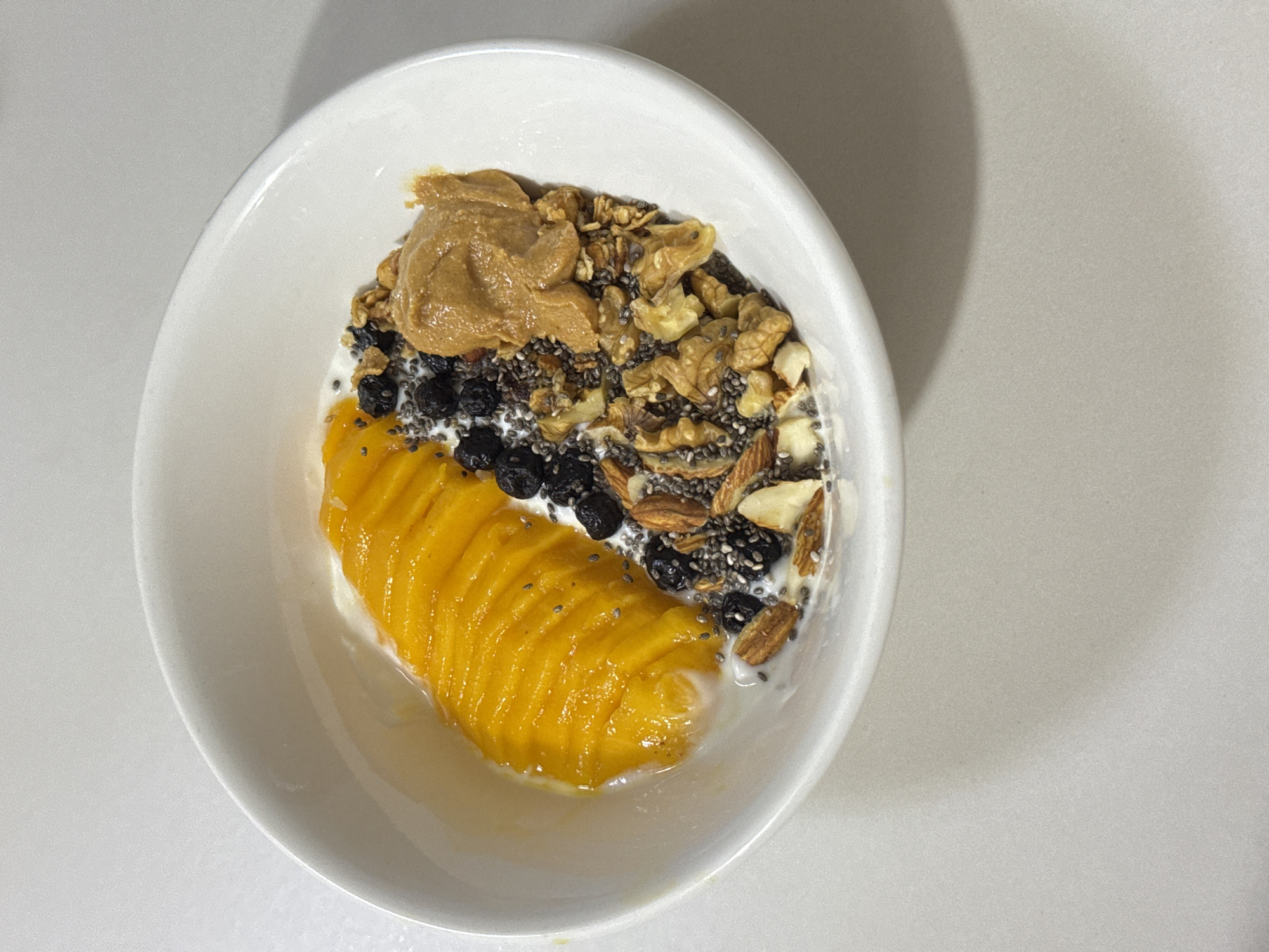 A close-up, overhead shot shows a white ceramic bowl filled with a healthy breakfast. The bowl contains a serving of yogurt, topped with thinly sliced mango arranged in a fan shape. Scattered around the mango are blueberries, chia seeds, and a mixture of nuts including almonds and walnuts. A dollop of peanut butter sits atop the nuts. The bowl is placed on a light-colored, textured surface, likely a countertop or table. The lighting creates soft shadows, suggesting an indoor setting. The scene is devoid of any identifiable landmarks or indicators of a specific location within Davao City, Philippines.