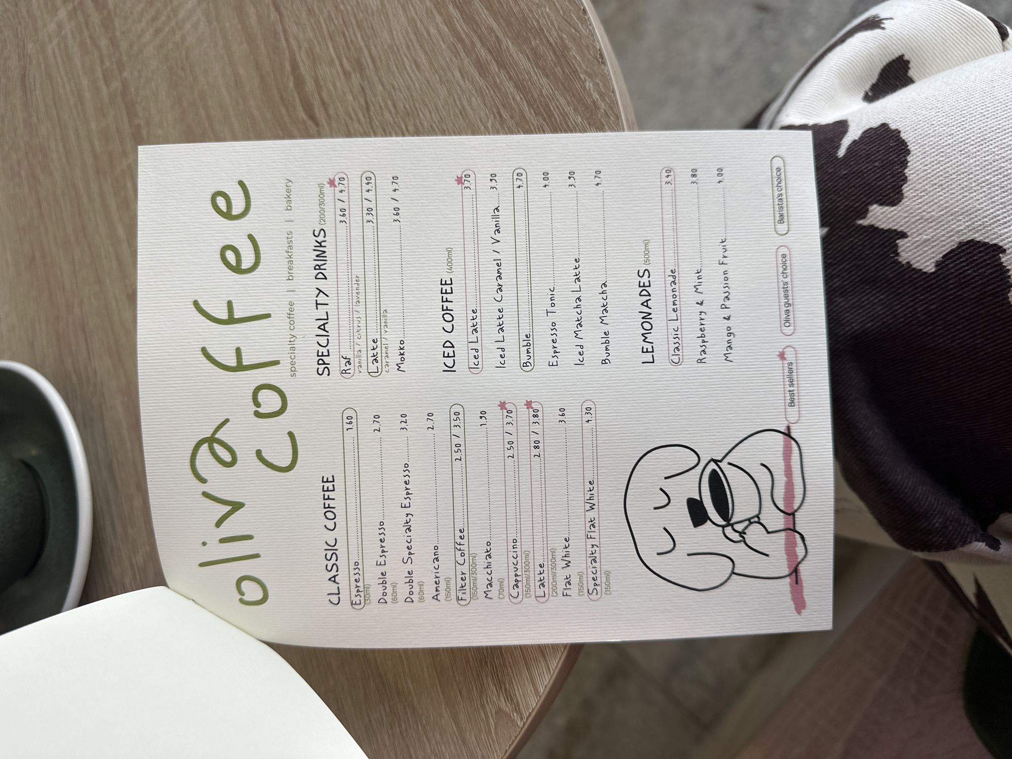 A top-down, slightly rotated, medium shot of a menu, primarily white with green text, on a light brown surface, likely a table. The menu is open, displaying a list of coffees and lemonades from "oliva coffee." On the left side is a dark green shoe, and on the right is a blurry patterned cushion. The menu lists various coffee drinks and lemonades with their corresponding prices in euro, with "classic coffee", "specialty drinks", "iced coffee", and "lemonades" as the categories. The menu also features a simple line drawing of a dog drinking from a cup at the bottom.
The location in the city of Šušanj, Montenegro, is not explicitly stated in the image, but the menu and objects suggest a cafe setting.