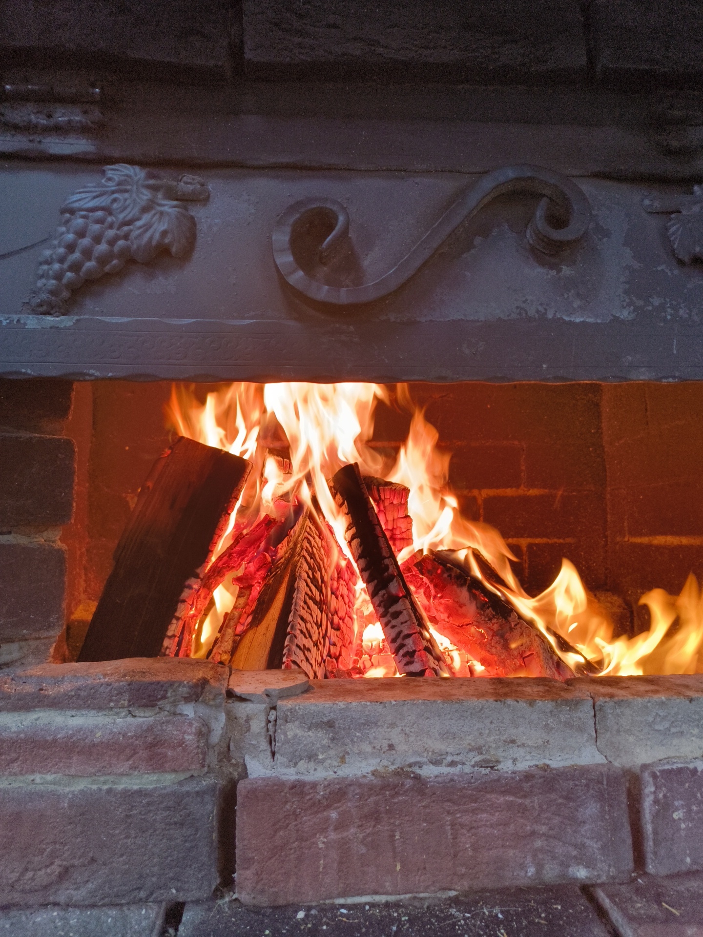The image shows a close-up view of a brick fireplace with a fire burning inside. The fireplace is decorated with a metal ornament depicting a bunch of grapes on the left and a swirl on the right. Logs are stacked and burning, producing bright orange flames and glowing embers. The bricks of the fireplace appear old and worn, with some cracks visible. The lighting is warm and dramatic, highlighting the intensity of the fire. There are no people or animals present in the image. The location is described as Востец Снт, Russia.