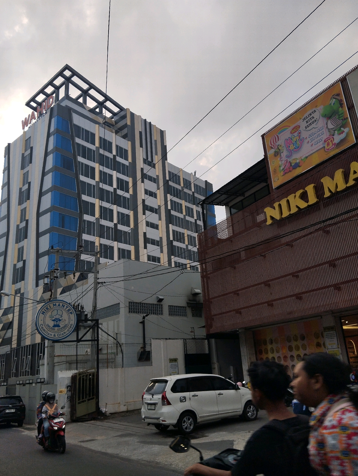 The image captures a street scene in Salatiga, Indonesia, under an overcast sky. Dominating the left side of the frame is the "WAHID" hotel, a modern, multi-story building with a distinctive geometric facade featuring alternating vertical panels of grey, white, and blue glass. A striking black lattice structure crowns the hotel. To the right, a building with a corrugated brown metal exterior displays a large, brightly colored billboard advertising "Mini Jelly" with a cartoon dinosaur mascot. Above this, yellow lettering spells out "NIKI MA". In the foreground, a white SUV is parked on the right side of the street. On the left, a motorcycle carries two passengers, a woman driving and another person behind her, as they proceed down the road. A circular blue sign for "NIKI MANTEP" is visible on a pole near the hotel. The street itself is paved, and various electrical wires crisscross the sky.