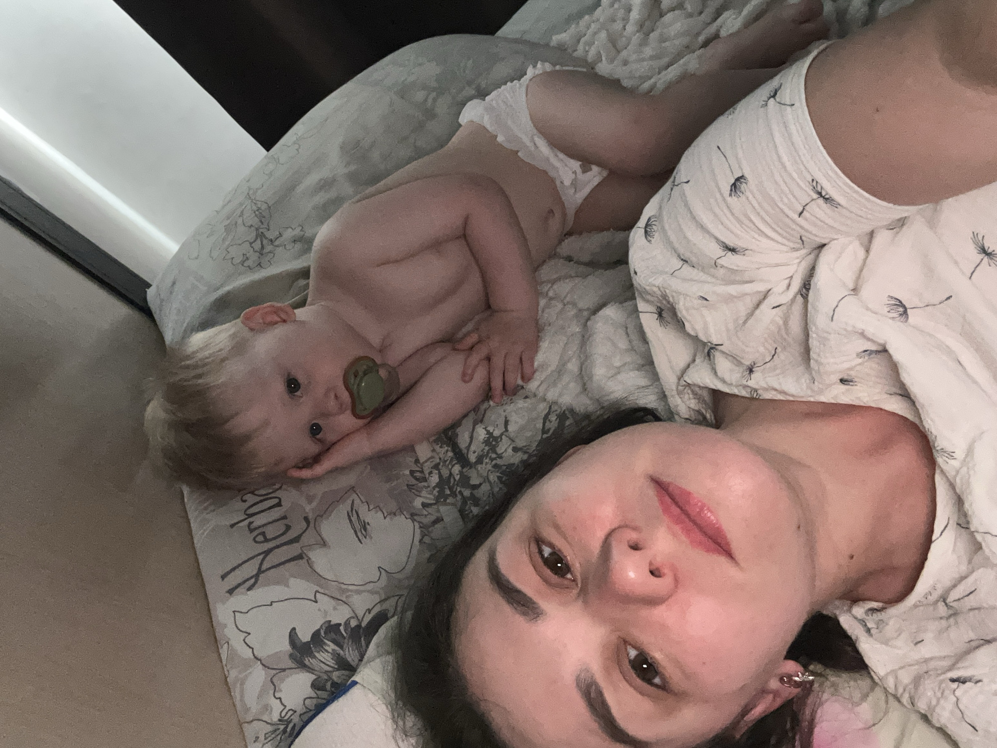 A woman and a baby lie on a bed in an indoor setting. The woman, with dark hair, brown eyes, and wearing lip gloss, is positioned in the foreground, facing the camera. The baby, with light hair and a pacifier in their mouth, is lying near the woman, wearing white clothing. The bed is covered with patterned bedding. The location is potentially within a dwelling in the city of Chaikovsky, Russia. The scene appears to be a candid moment between the woman and the baby.