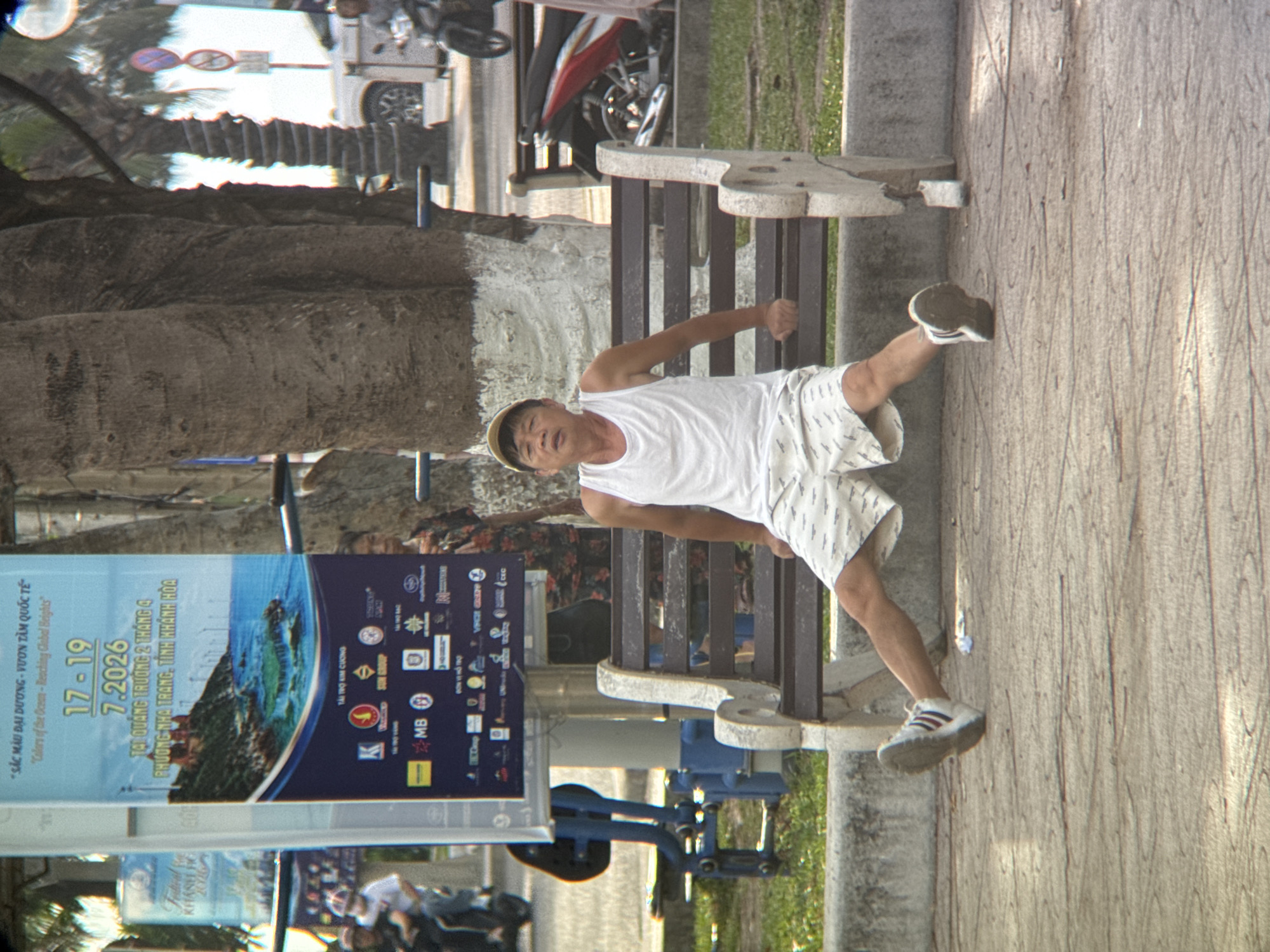 A light-skinned man wearing a white tank top and patterned white shorts with a yellow cap is sitting on a dark wooden bench with concrete legs. He is looking towards the camera with his mouth slightly open and his hands resting on the bench. His left leg is extended, and his right leg is bent at the knee with his foot on the ground. The bench is on a patterned concrete walkway, with grass and palm trees in the background. To the left of the bench is a blue and grey exercise machine. Behind the man is a large banner with Vietnamese text and images of the ocean and buildings. The banner advertises an event in Nha Trang, Vietnam from July 17-19, 2026. To the right of the bench is a parked red and black motorcycle and a person walking away from the camera. The lighting suggests it is daytime.