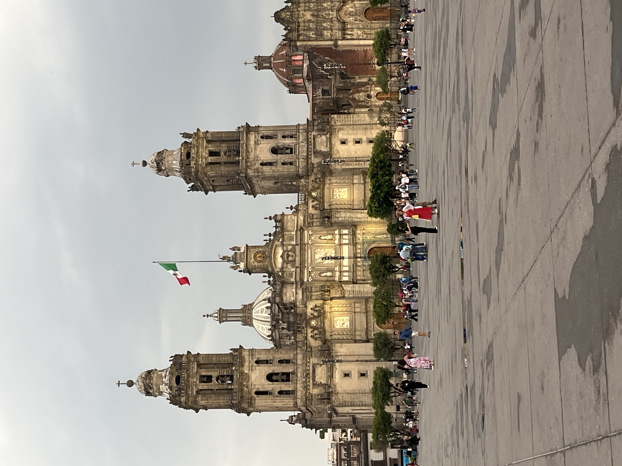 The image shows the Metropolitan Cathedral in Mexico City, Mexico. The cathedral is a large, ornate structure with multiple towers, domes, and intricate carvings. The Mexican flag is flying atop one of the towers. In front of the cathedral is a large paved square with many people walking and standing around. The sky is overcast.