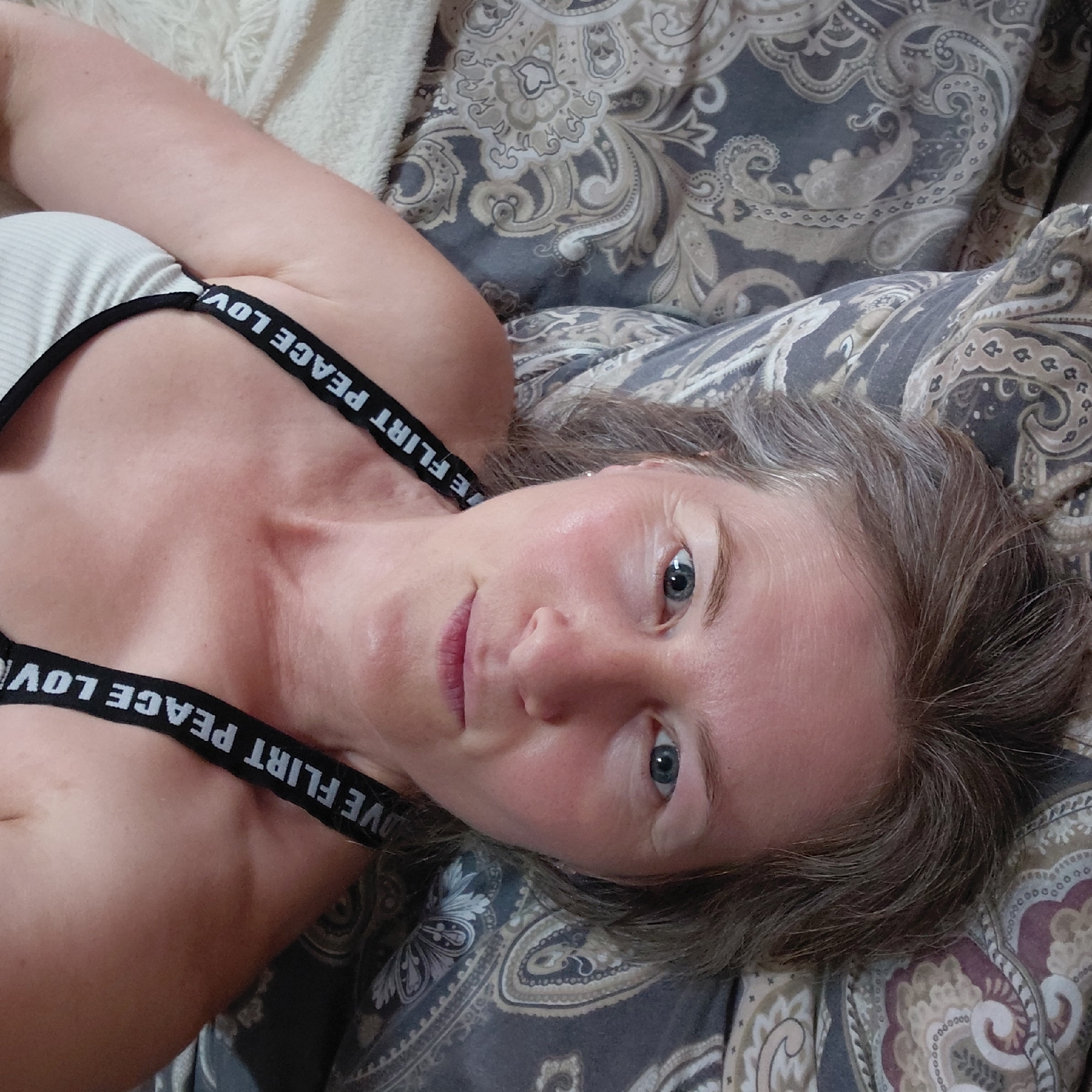 The image shows a close-up, overhead view of a woman lying on a bed, facing upwards towards the camera. She has graying brown hair that is slightly disheveled, and her face is illuminated by soft lighting. Her eyes are blue, and she has faint wrinkles around them. She is wearing a white ribbed tank top with black straps that have white text printed on them, reading "LOVE FLIRT PEACE LOVE". The background consists of a patterned duvet with paisley designs in shades of grey, beige, and deep red. To the left of the woman, a white fuzzy blanket or rug is visible. The scene appears to be an indoor setting, likely a bedroom, with no identifiable landmarks of Novosibirsk, Russia.