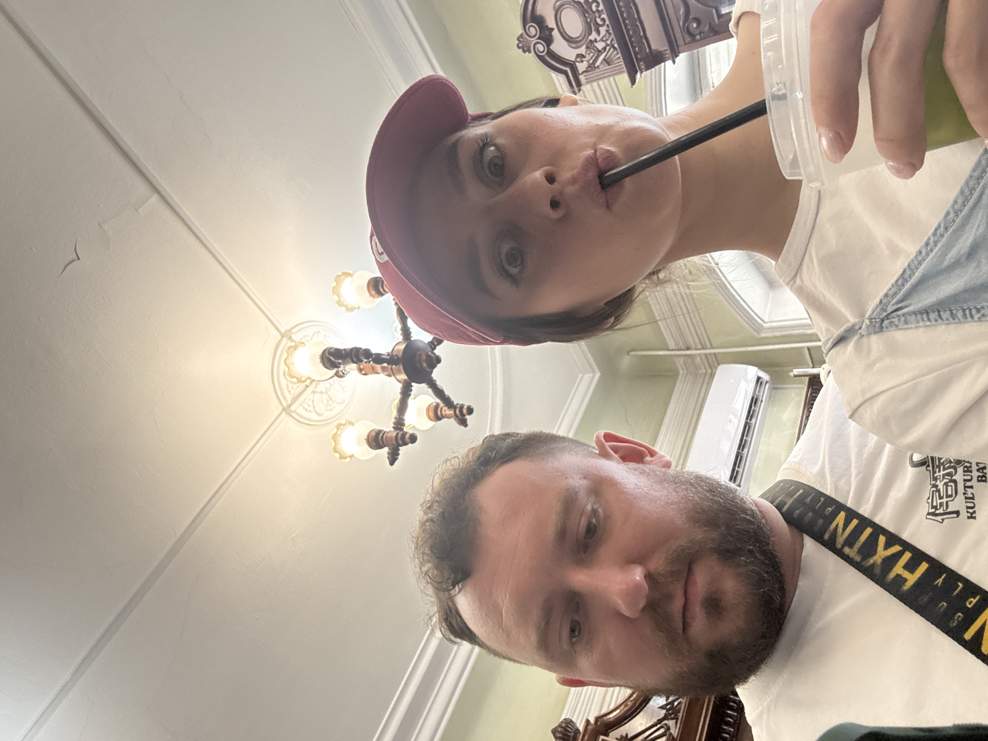 A selfie taken from below depicts two people indoors, likely in a building in Krasnoyarsk, Russia. A woman in a red baseball cap is holding a drink with a straw to her lips. A man with a beard is positioned below her. An ornate chandelier is visible near the woman's face. An air conditioner is on the wall in the background. A decorative wooden fixture is on the bottom side of the frame. The man is wearing a white T-shirt with a black and yellow strap.