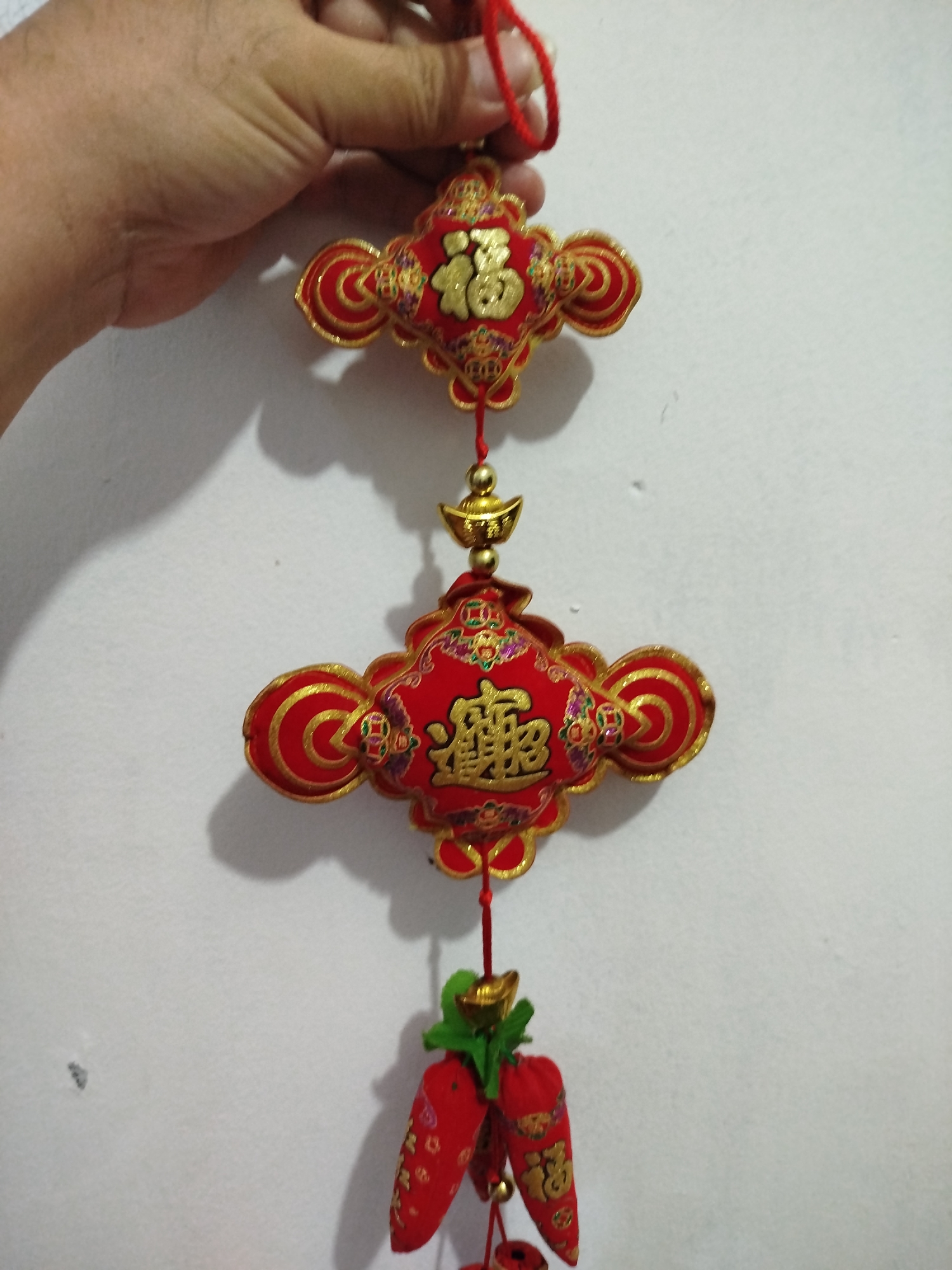 A hand holds a red and gold Chinese New Year ornament featuring two decorative knots with gold Chinese characters, connected by a gold bead and an inverted gold ingot. Below the lower knot are three small red fabric carrot-shaped charms adorned with gold Chinese characters and small gold bells. The ornament is hanging against a white wall. The image is cropped closely to the ornament and the hand, with the background being a plain white wall. There is no discernible information about the location in Bandungrejo, Indonesia.