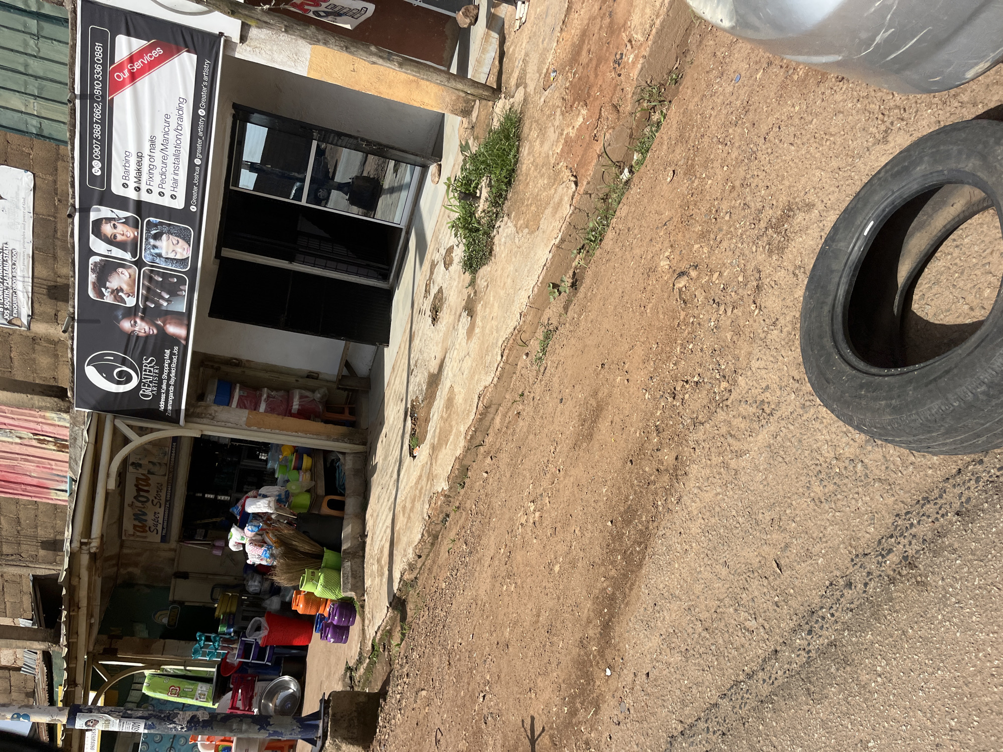 The image depicts a street scene in Gura Top, Nigeria. In the foreground, a worn tire lies on a dirt and asphalt road. To the right, the silver fender of a vehicle is visible. The background reveals a row of shops. One shop, "Fantora Super Stores," displays various household items such as buckets and brooms. Adjacent to it is a salon with a prominent banner advertising "GREATER'S ARTISTRY," listing services like "Barbing, Makeup, Fixing of nails, Pedicure/Manicure, and Hair installation/braiding." The banner also shows contact information and social media handles. The shops are built close together, with visible corrugated metal roofing and concrete facades. The ground in front of the shops is paved with concrete, showing signs of wear and tear, with some vegetation growing through cracks. The overall impression is of a bustling commercial area with a mix of retail and service businesses.
