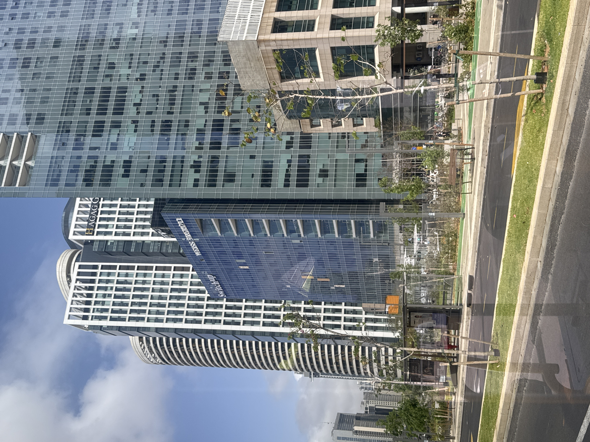 The image depicts a street in Tel Aviv, Israel, featuring modern architecture. On the left, several skyscrapers are visible, including buildings labeled "AGAG" and "Swissdigital." The buildings showcase a contemporary design with glass facades. The sky is partly cloudy. On the right side of the image, a street with a sidewalk and trees is present. A bus stop with advertising is located along the sidewalk. The overall scene conveys an urban environment within a modern city.