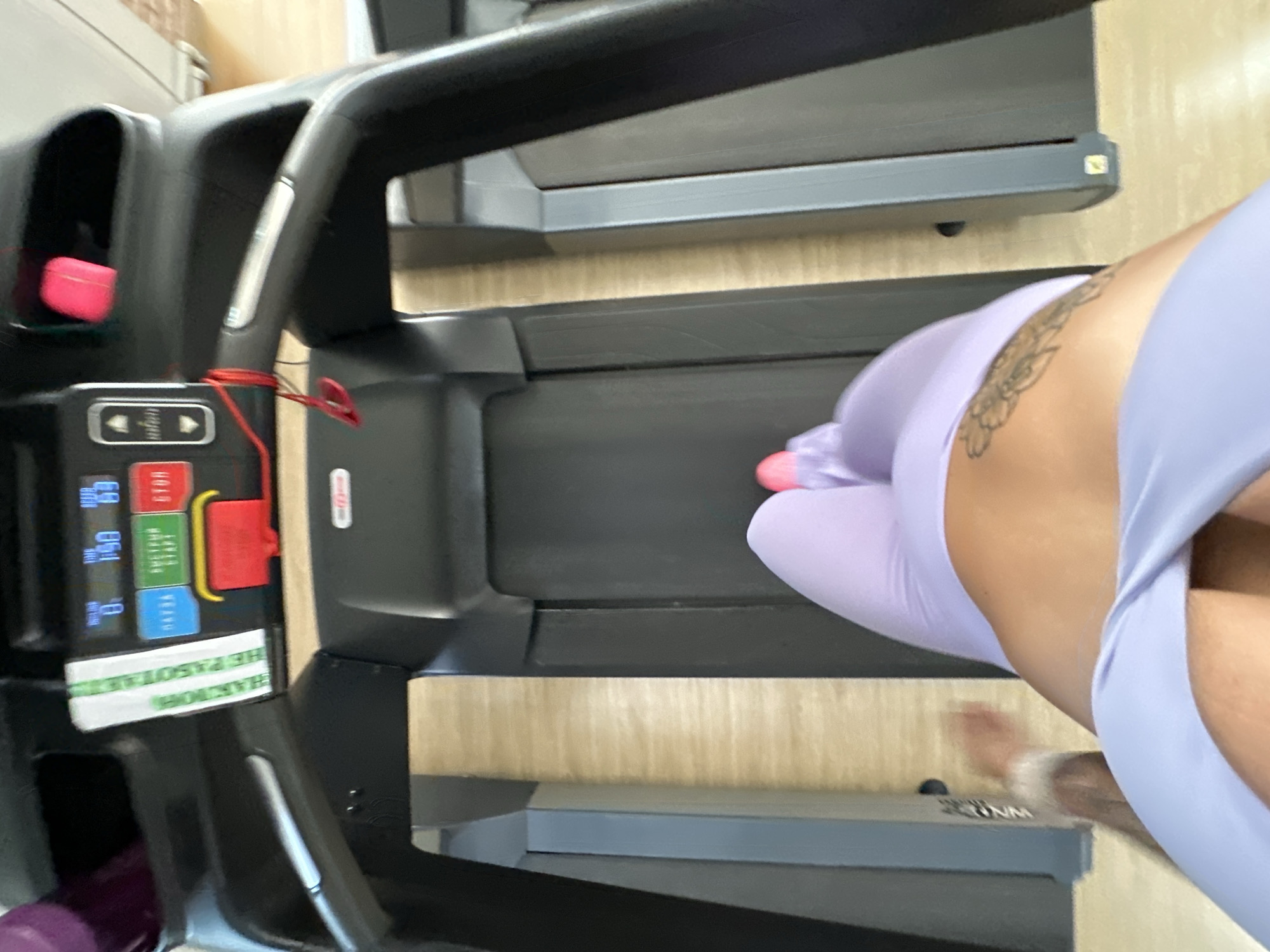 The image shows a person on a treadmill. The person is wearing light purple leggings and a matching sports bra. They have a tattoo on their hip. The treadmill is in a gym, with other treadmills visible in the background. The floor is light brown. The image is taken from a high angle, looking down at the person and the treadmill. The scene is well-lit, with natural light coming from the right side of the image. It is not possible to determine the specific location within Yuzhnouralsk, Russia.
