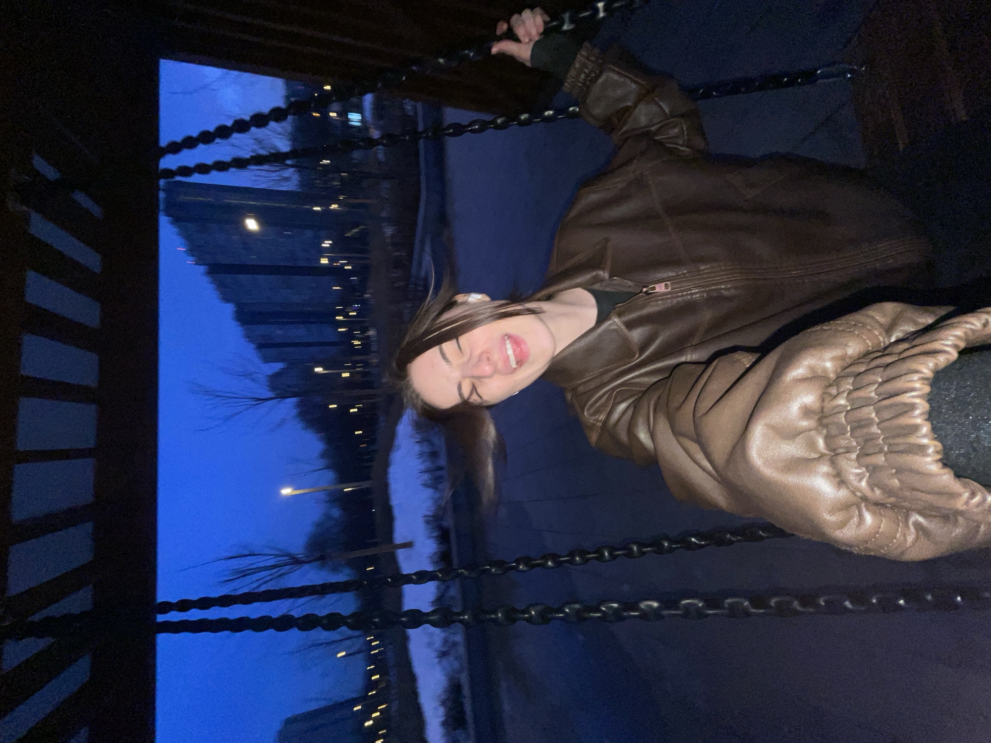A young female is depicted in an outdoor park setting, situated on a swing during twilight or night. Her face, partially obscured by wind-blown hair, is directed towards the camera with eyes squeezed shut, mouth open in an expression of laughter or exhilaration, and tongue slightly visible. She wears a brown leather jacket, dark undershirt, and a white wireless earbud in her right ear. Both hands grip the dark metal chains of the swing. The foreground consists of dark ground, while patches of snow are visible in the midground, interspersed with bare trees and numerous illuminated path lights. In the background, distinctive multi-story residential buildings, with a stepped or angular profile and visible internal lighting, rise against the deep blue sky. This scene is consistent with a modern urban park in Moscow, Russia.
