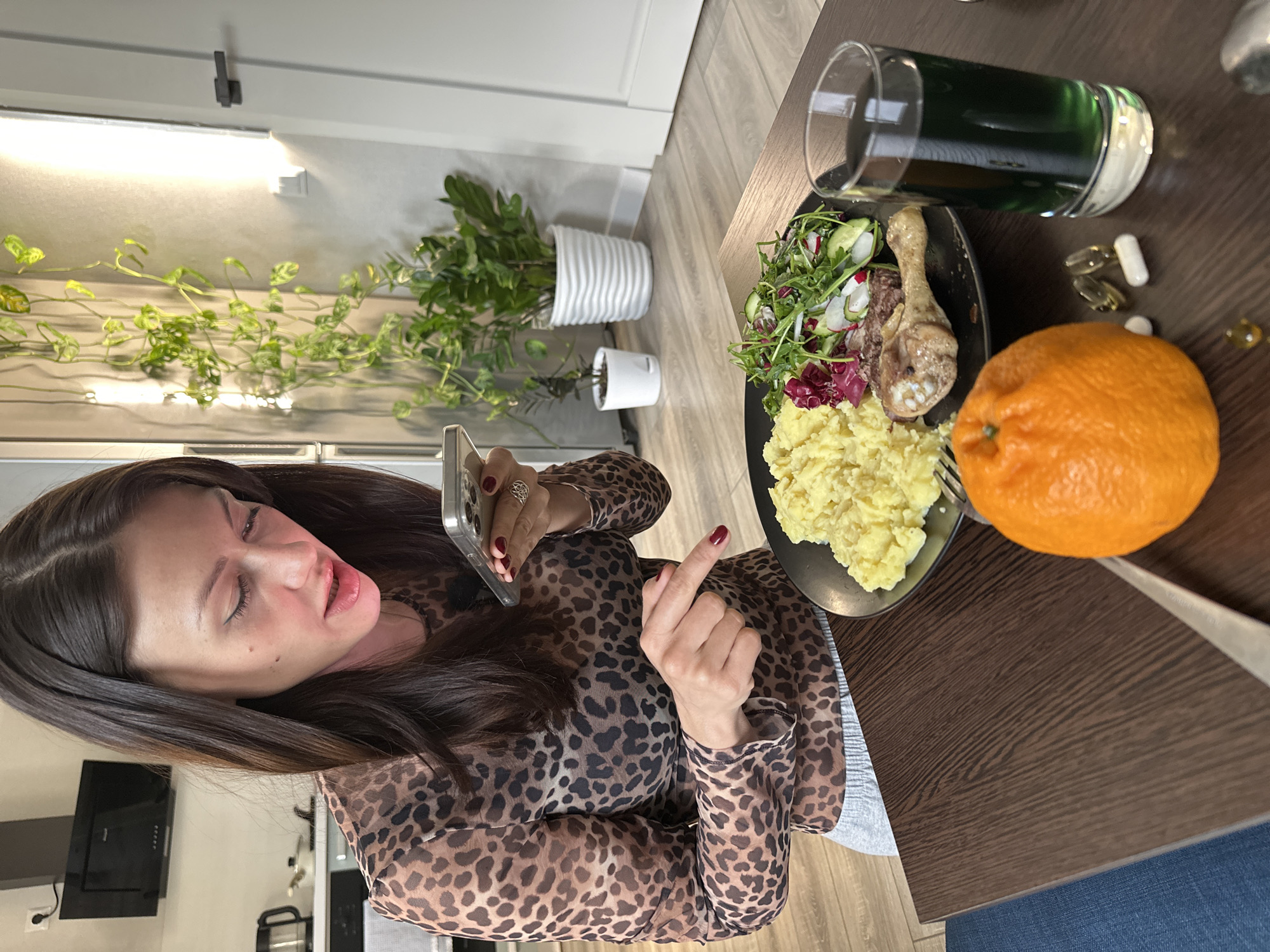 The media file captures an indoor scene, likely a modern apartment in Murino, Russia, focusing on a woman and a meal laid out on a dark wooden table.
The main subject is a woman with long dark hair, positioned on the left side of the frame. She is wearing a leopard print long-sleeved top. Her left hand holds a smartphone, while her right hand is raised, with her index finger pointing forward. She has red nail polish on both hands. Her eyes are partially closed, and her mouth is slightly open, suggesting she might be speaking or reacting to something.
On the table in front of her is a meal consisting of a plate with mashed potatoes, a piece of cooked poultry (possibly a chicken leg), and a salad featuring mixed greens, radicchio, and sliced radishes or cucumbers. Alongside the plate, there's a whole orange. A glass filled with a dark green liquid (possibly a supplement drink or smoothie) is also on the table, next to which are several pills and capsules, including clear gel capsules and white capsules.
The background shows light wooden flooring and white walls. Several potted plants are visible, including a larger plant in a white ribbed pot and a smaller one in a white pot. A climbing plant is also seen along a wall, near a linear light fixture. A black range hood can be faintly observed in the far left background, indicating a kitchen area. The overall lighting suggests it is daytime or a well-lit interior. No visible text is discernible in the image.