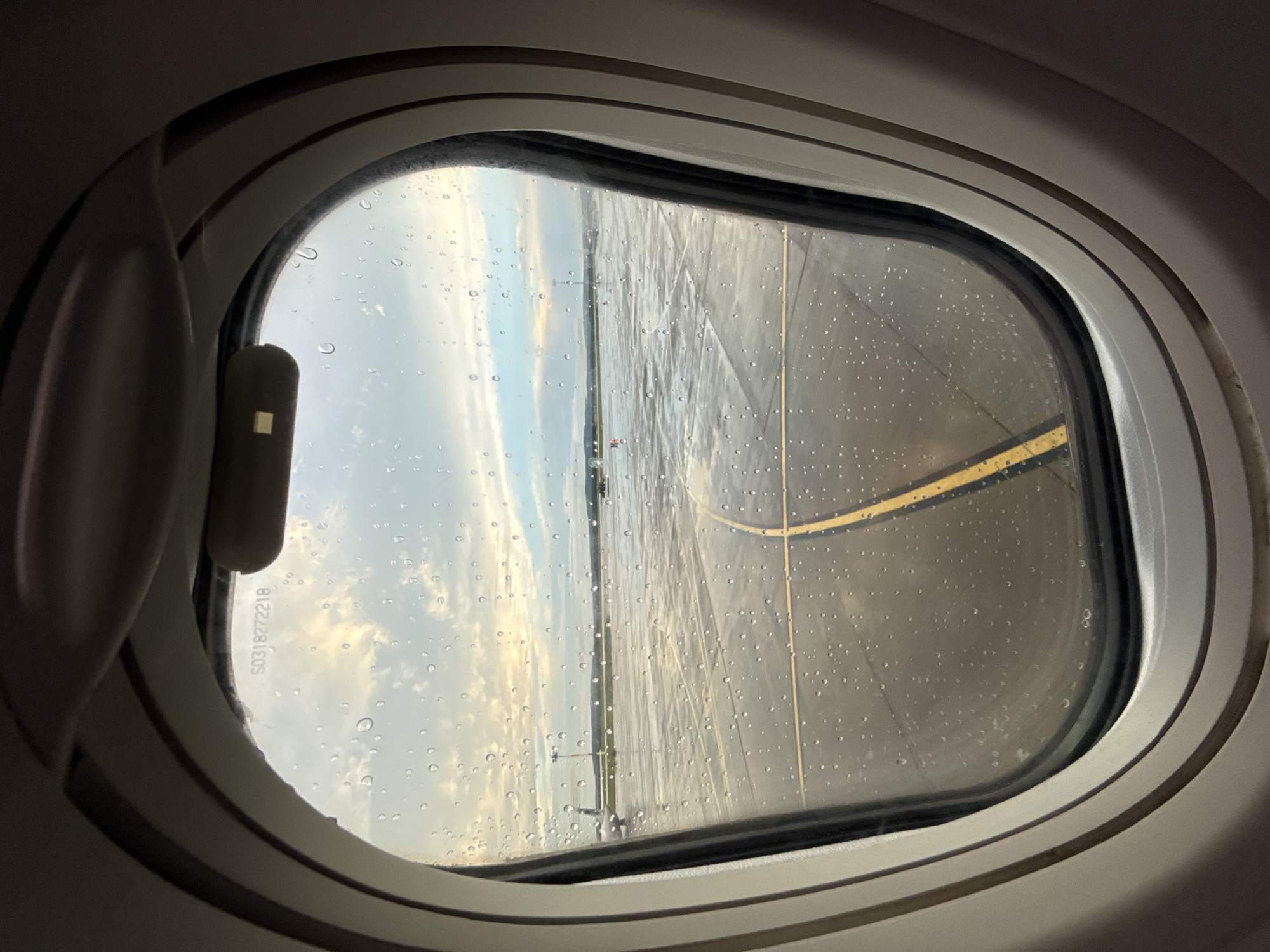 The image is taken from inside an airplane window, looking out at an airport tarmac. The window has numerous raindrops on its surface, obscuring the view slightly. Outside, the tarmac is wet, reflecting the sky, which is overcast with patches of sunlight breaking through. Yellow lines mark the taxiway. In the distance, there are airport lights, a control tower, and what appears to be another aircraft. The scene is set at Koltsovo International Airport (SVX) in Yekaterinburg, Russia. There are no people visible in the image.