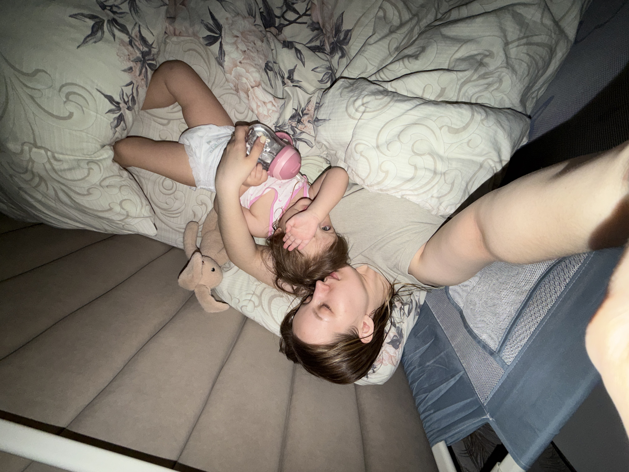 The image depicts a woman lying on a bed with a young child. The woman is on her back, with her head resting on a pillow with a floral pattern. The child is lying on top of her, holding a bottle with a pink lid. The child's hand is partially covering their face. A stuffed animal is next to the woman's head. The bed is covered in a floral patterned duvet. The location is not discernable from the provided image.