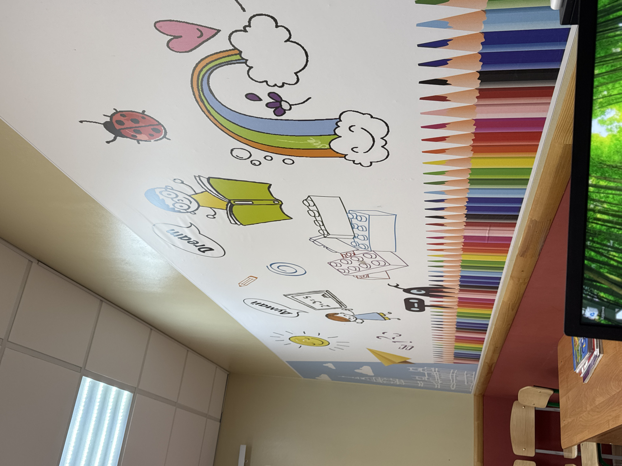 This image captures an interior view of a vibrant children's space, likely a classroom or playroom, adorned with colorful, cartoon-style illustrations on a wall or ceiling. The perspective looks upwards, revealing a bright white surface covered with engaging drawings.
Key illustrations include a red ladybug, a rainbow arching between two fluffy clouds, accompanied by a pink heart and a small purple flower. A prominent border of numerous colorful pencils, arranged side-by-side in a full spectrum of hues, lines the right edge of the decorated surface. Other drawings depict an open green book, a spiral notebook, various stationery items like a loose pencil, and building blocks resembling LEGOs. A smiling yellow sun, a paper airplane, and a drawing of a child standing by a blackboard with "2+3=5" written on it are also visible. Another cartoon figure appears to be reading, with the word "Dream" written nearby in a playful font. The word "Duygu" is also seen near a pencil illustration and a swirl.
To the right, a dark monitor screen displays a green, possibly nature-themed, background. Below it, parts of wooden, child-sized tables and chairs are visible against a reddish-brown wall. On the left, a wall featuring white square panels includes a bright rectangular fluorescent light fixture. The surrounding walls are a light beige color, complementing the cheerful decorations. The overall atmosphere is bright and inviting, designed to stimulate creativity and learning in children. The location context is Perm, Russia.
