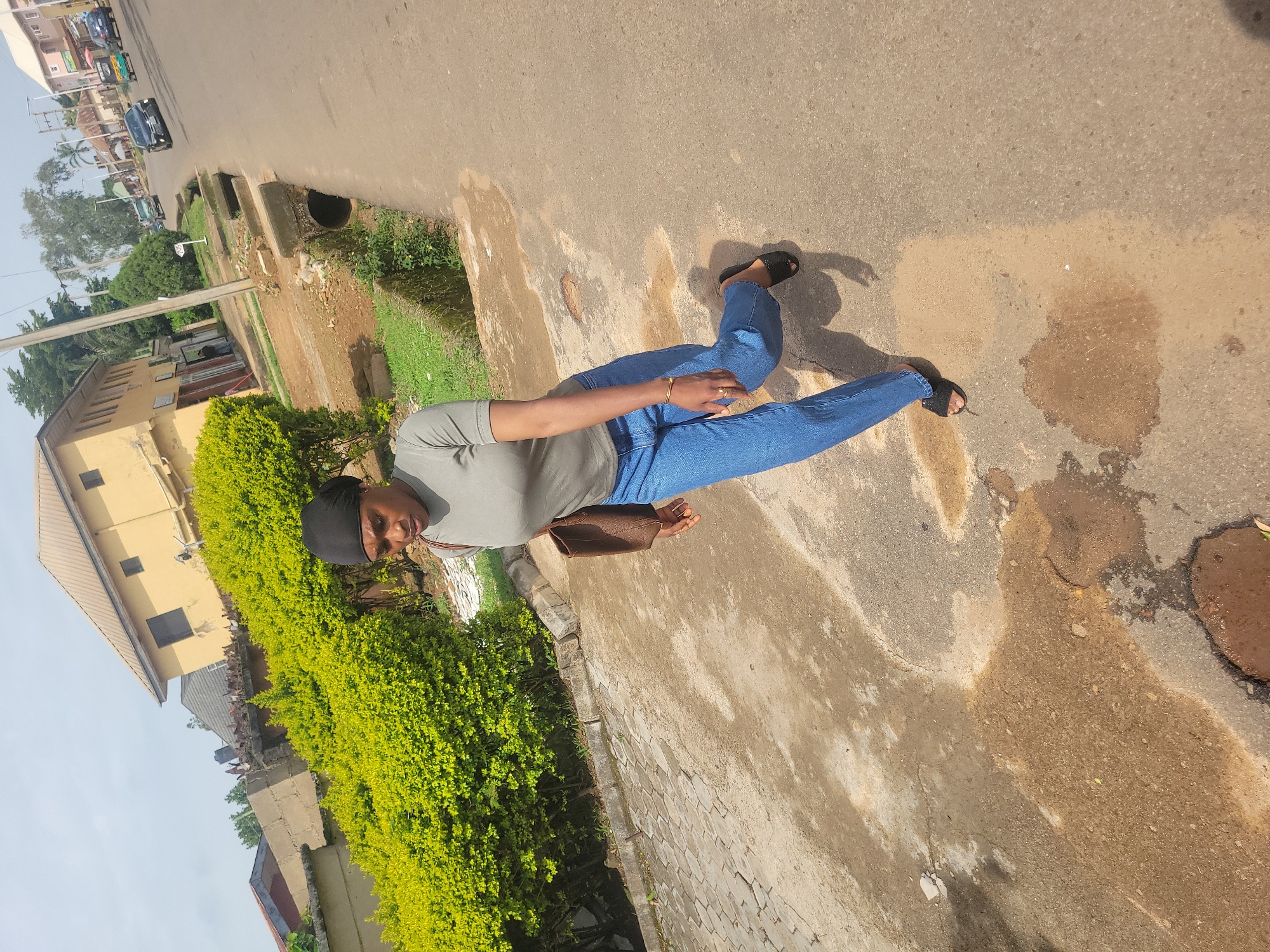 A young woman with a dark headscarf and a grey t-shirt walks on a paved path next to a road in Jos, Nigeria. She is wearing blue jeans and black slip-on sandals, and carrying a brown tote bag on her right shoulder. The path has some puddles of water and debris. In the background, there is a yellow building with shops on the ground floor, a drainage channel, and a street with vehicles. The sky is clear and bright.