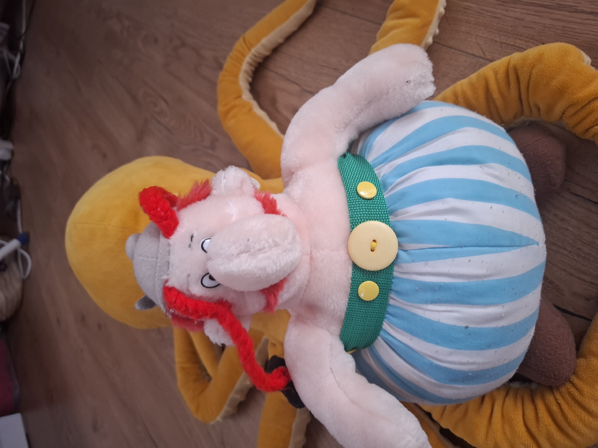 The image shows a close-up, eye-level shot of a plush toy of the comic book character Asterix. Asterix is wearing his characteristic blue and white striped skirt, a green belt with two yellow buttons, and a grey helmet with red hair. He is propped up against a large, yellow plush octopus, whose tentacles are spread out around him. The toys are lying on a wooden floor, which appears to be in a domestic setting. The lighting is natural, coming from the top right of the frame. The overall impression is one of a casual, indoor scene. There is no indication of the specific location within Montreuil, France, or any other contextual information that would allow for a description of the city.