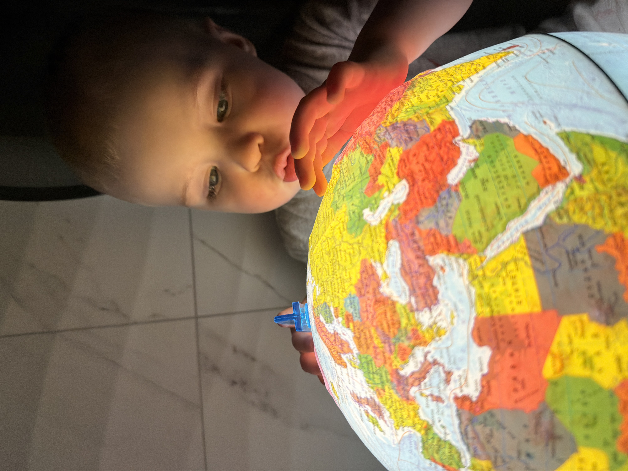 A child is inspecting a glowing globe. The globe shows land masses colored in yellow, green, red, and brown. The child has fair skin, light brown hair, and is wearing a grey shirt. Their hand is illuminated by the globe's light. The background is dim, revealing a tiled floor. This image was not taken in the city of Votkinsk, Russia.
