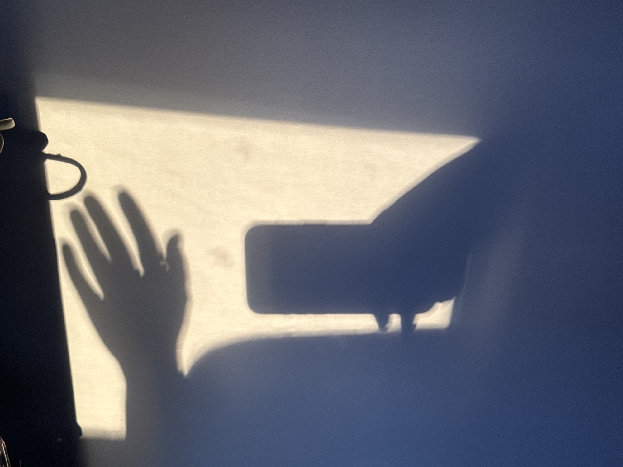 The image captures a scene dominated by strong shadows cast upon a textured, dual-toned surface, likely a wall or floor. The primary subjects are the distinct shadows of a human hand and a smartphone. The hand, visible on the left side, shows clear finger outlines, with a prominent ring casting its own shadow on one finger. It appears to be holding the rectangular shape of a smartphone, whose shadow extends to the right from the hand. This arrangement suggests the activity of someone taking a picture or video, with their hand and phone positioned to block a light source.
To the far left, a partial shadow of an object with a D-shaped handle is visible, possibly a mug or a lamp fixture. The scene is brightly lit by what appears to be natural light, most likely sunlight, entering from the left. This light creates a warm, yellow-white hue on the illuminated part of the surface, contrasting sharply with the darker, bluish-grey area not reached by the direct light. The distinct edges and elongation of the shadows imply a strong, low-angle light source, characteristic of morning or late afternoon. There is no visible text in the image.