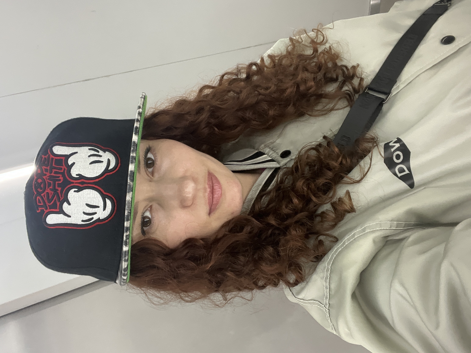 A single female individual is depicted in a head-and-shoulders portrait, centered in the frame. She possesses long, curly, reddish-brown hair and brown eyes, looking directly at the viewer with a neutral to slightly smiling expression. She is wearing a black flat-brim baseball cap with prominent red embroidered text "DOPE SHIT" above two stylized white cartoon hands, reminiscent of Mickey Mouse gloves, making a "rock on" gesture. The cap's brim has a green edge and a black and white patterned underside. Her attire includes a light grey or off-white bomber-style jacket featuring a ribbed collar with black and white stripes, black snap buttons down the front, and visible white contrast stitching on the shoulder. A black trapezoidal patch with the white text "DOW" is present on the lower left chest of the jacket. A black fabric strap, consistent with a shoulder bag, runs diagonally across her chest. The background is a plain, light-colored wall or partition, suggesting an indoor setting within Saint Petersburg, Russia. Faint vertical lines or seams are visible on the light grey wall behind her.