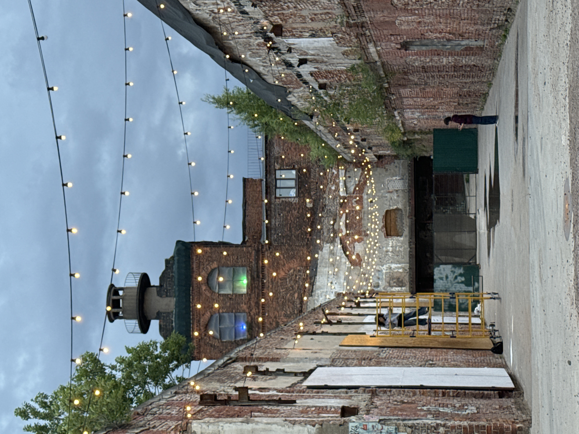 The image depicts an outdoor courtyard area with weathered brick buildings on either side, suggesting an industrial or historic setting. String lights are draped across the space, illuminating the scene against an overcast sky. In the background, a tower-like structure rises above the buildings, resembling a lighthouse or industrial chimney. Two individuals are visible in the courtyard: one on a yellow scaffold on the left, appearing to be working or posing, and another standing further back on the right, looking at a phone. The ground is paved and shows signs of wear and some puddles, indicating it is likely an urban environment. The overall impression is one of a revitalized industrial space or an artistic venue in Saint Petersburg, Russia, possibly a former factory or port area.