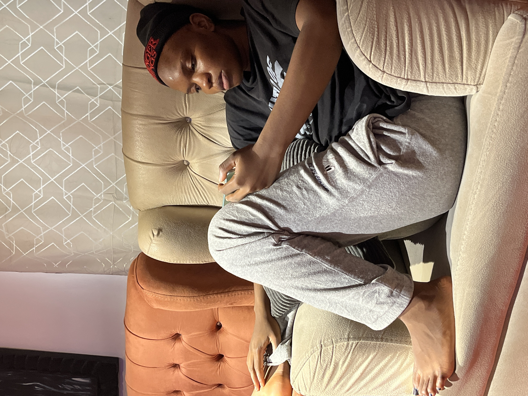 The image depicts a young Black person seated on a tufted sofa in a domestic indoor environment. The person is wearing a black t-shirt and grey sweatpants, with a black beanie adorned with red lettering. They are holding a mobile phone in their hands and appear to be engaged with it. The sofa is upholstered in a beige fabric with tufted accents, and there is an orange tufted sofa segment to the left of the frame. The background features a light-colored curtain with a geometric pattern. The lighting suggests an evening setting. The person's bare feet are visible, resting on the sofa. There is no indication of the specific location within the city of Jos, Nigeria, beyond it being an indoor residential setting.