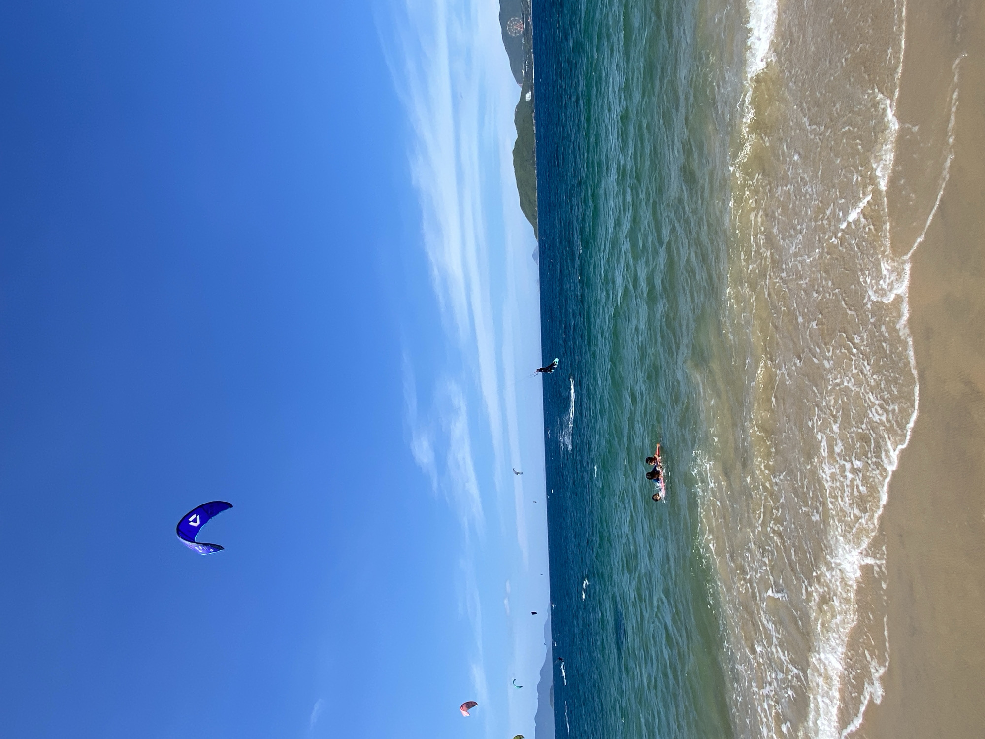 This image captures a vibrant scene at a beach in Nha Trang, Vietnam. The sky is a brilliant, clear blue, dotted with wispy white clouds. In the upper left quadrant, a large, blue kite with a white logo soars against the sky. The sea is a deep, rich blue, transitioning to turquoise closer to the shore. Gentle waves with white foam lap at a sandy beach in the foreground. Several individuals are engaged in water sports. In the middle ground, three people are floating in the water, appearing relaxed. Further out, multiple kitesurfers are actively riding the waves, their colorful kites dotting the horizon. In the far distance, a lush green island with a Ferris wheel visible on its side sits against the backdrop of a hazy coastline. The scene conveys a sense of active recreation and enjoyment of a sunny day at the beach.