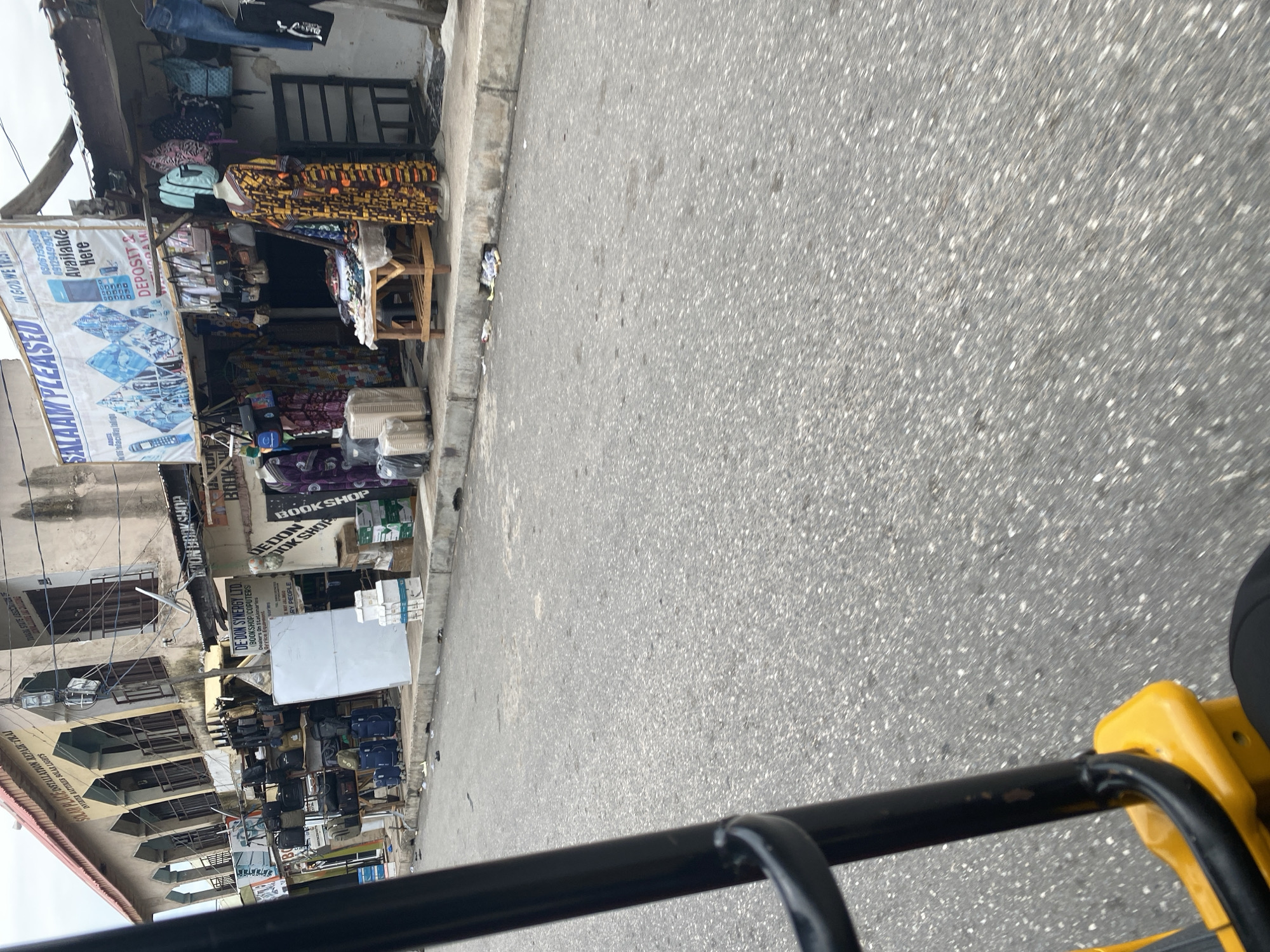 The media file shows a street-level view of a commercial area in Jalingo, Nigeria. The scene is dominated by various small shops and stalls lining a paved road. One prominent shop, "DE-DON SYNERGY LTD," appears to be a bookshop and computer store, selling stationery, office equipment, and accessories. Another stall displays colorful fabrics and clothing on a mannequin and on racks. Bags of various sizes and types are also for sale. Signs indicate services like "SOLAR PLACE INSTALLATION REPAIR TRAJ" and "INVERTER BATTERIES SOLAR LIGHTS." A banner reads "SALAAM PLEASED IN GOLWE" with contact numbers. The setting suggests a busy marketplace or street with multiple vendors. The lighting indicates it is daytime. The perspective suggests the photo was taken from a moving vehicle or slightly elevated position, as evidenced by the blurred asphalt in the foreground and the yellow frame of what might be a tricycle or similar vehicle. There are no people clearly visible in the image, but the presence of shops and goods implies commercial activity.