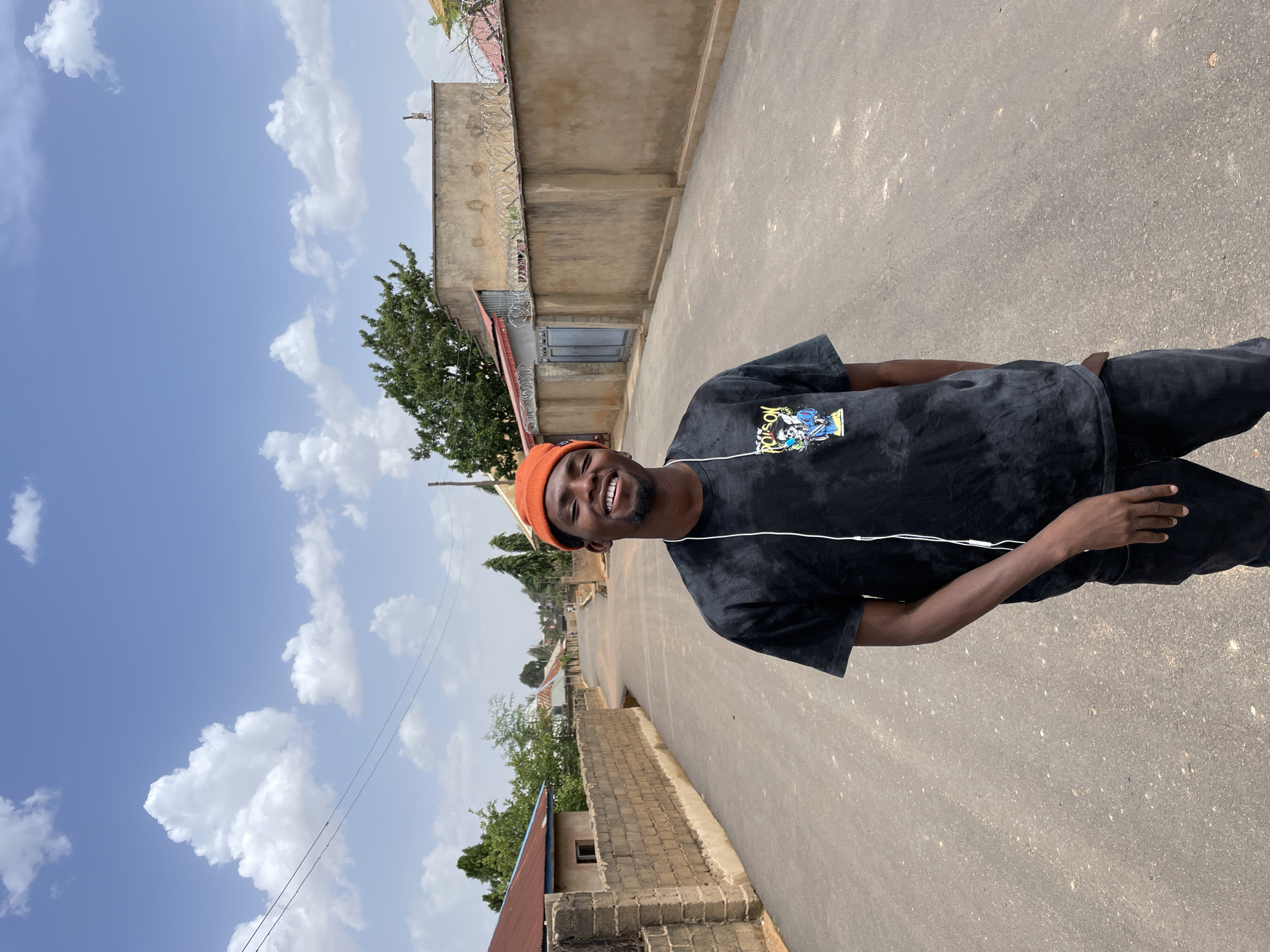 A young, dark-skinned man with a beard and an orange beanie stands on a paved road in Gura Top, Nigeria. He wears a tie-dye black t-shirt and dark pants, and has earbuds connected to his phone. He is smiling at the camera. Behind him are buildings, walls, and trees typical of the Nigerian landscape, set against a blue sky with scattered clouds.