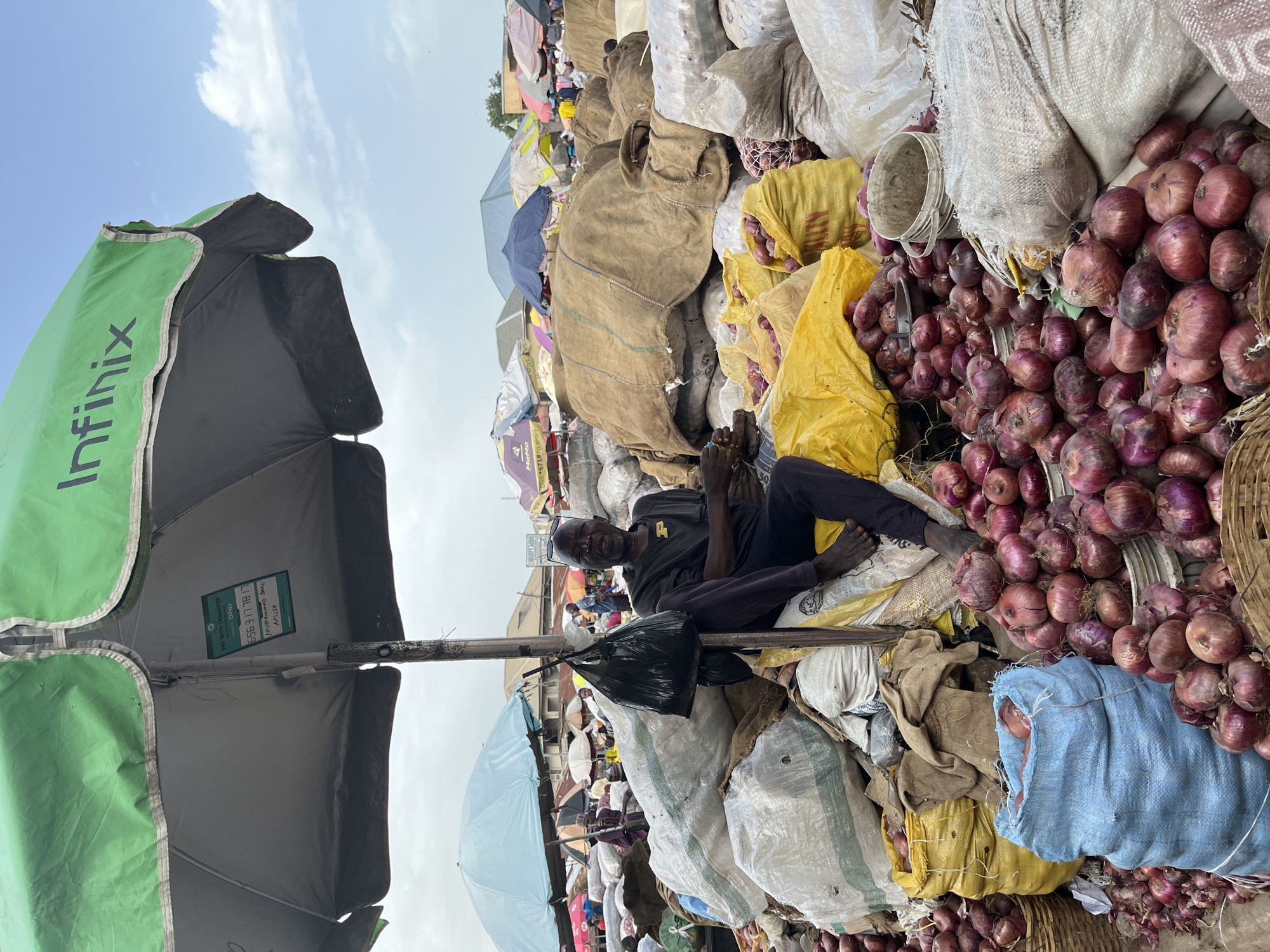 An overhead view captures a bustling outdoor market scene in Rafin Par, Nigeria. A prominent green Infinix-branded umbrella shades a man seated amidst piles of produce. The man, wearing a dark t-shirt and pants, appears to be a vendor, with his bare feet visible and his hands resting in his lap. He is surrounded by large sacks of goods, predominantly onions. Some sacks are made of rough burlap, while others are white or blue plastic. Several baskets overflow with red onions, and a bright yellow tarp is used to cover a portion of the goods. In the background, other market stalls and umbrellas are visible, along with a cloudy sky. A black plastic bag hangs from the umbrella's pole. A small sign attached to the umbrella pole displays text including "PAY WITH TRANSFER," "OPay," and a phone number. The overall impression is one of a vibrant, active marketplace.