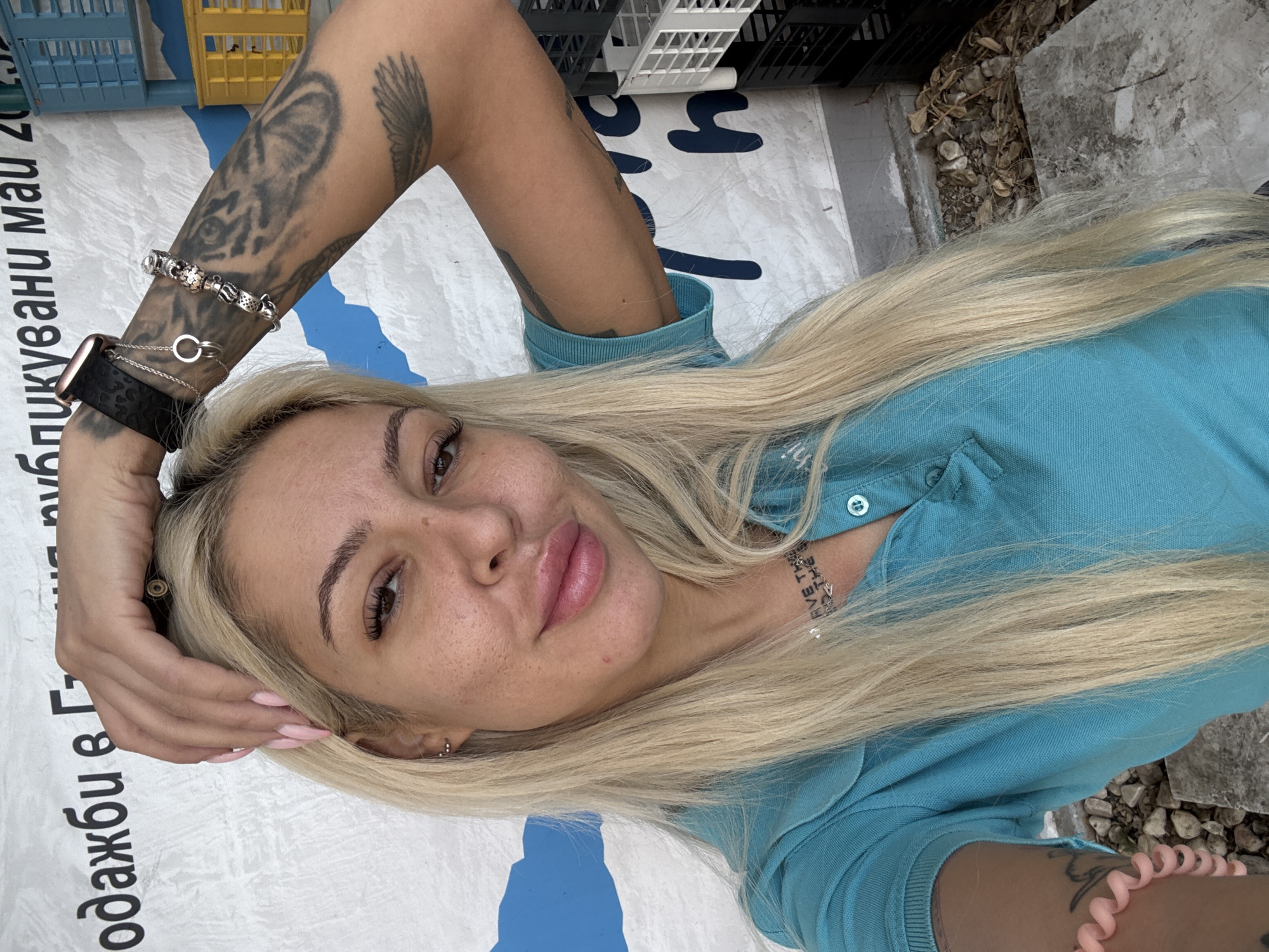 A woman with blonde hair and tattoos is lying down, possibly in Plovdiv, Bulgaria. She is wearing a light blue collared shirt and jewelry including an Apple Watch, bracelets, and a necklace. She is lying on a white surface with blue lettering in Cyrillic. Plastic crates are stacked above her head. The ground is partially visible, showing stones or gravel.