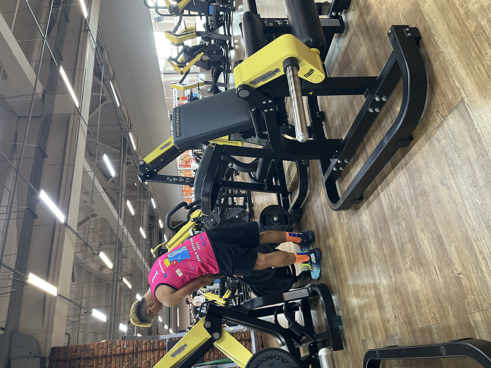 In a gymnasium located in Chaikovsky, Russia, a man with dyed blonde hair is standing next to a piece of exercise equipment. He is wearing a bright pink tank top with the number "1" on it, black shorts, and colorful athletic socks and shoes. The tank top has Cyrillic text and graphics. The man appears to be in the process of setting up or adjusting the weight machine. The gym has a wooden floor and industrial-style lighting. Other gym equipment, including yellow and black machines, are visible in the background, along with brickwork on one wall. The overall environment suggests a functional, modern fitness facility.
