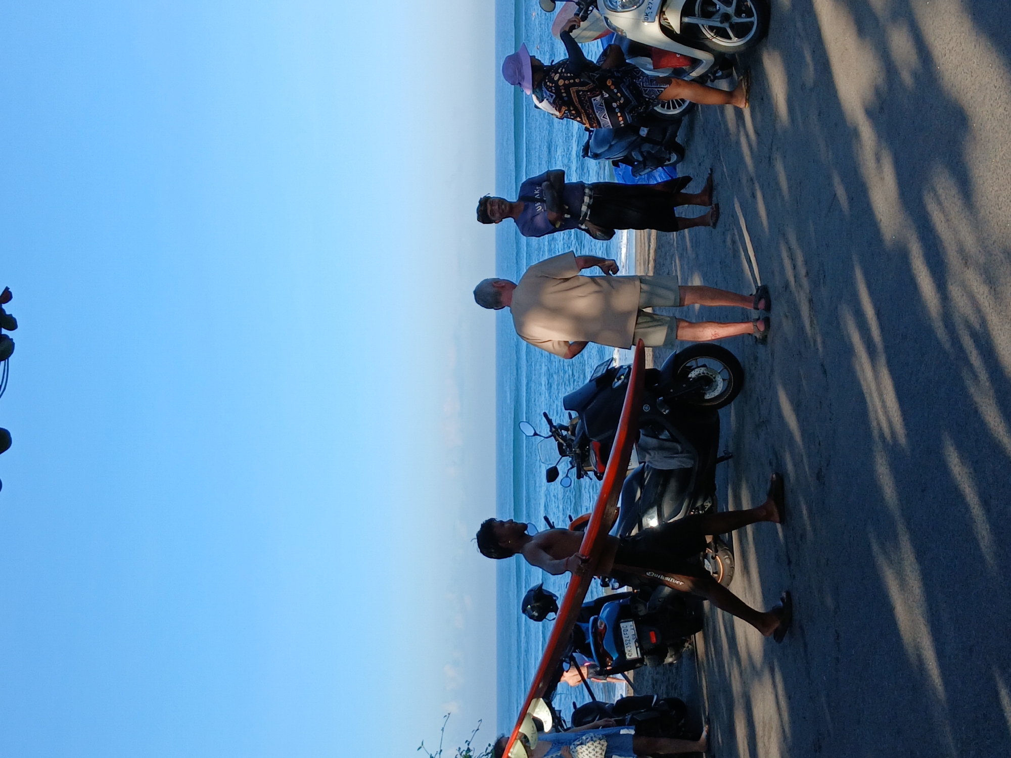 The image depicts a beach scene in North Kuta, Indonesia. In the foreground, several motorcycles are parked on a paved surface. A young man is walking, carrying a red surfboard on his right shoulder. He is shirtless and wearing dark shorts. To his left, a woman in a blue dress is standing near a motorcycle, holding a white bag. Further to the right, two men are talking. One man, wearing a tan shirt and shorts, is facing away from the camera. The other man, wearing a dark purple shirt and a sarong, is facing the camera with his arms crossed. Next to him, a woman wearing a patterned top and a purple hat is leaning on a white scooter. In the background, the ocean is visible with gentle waves, and the sky is clear and blue. The image captures a casual moment on the Indonesian coast.