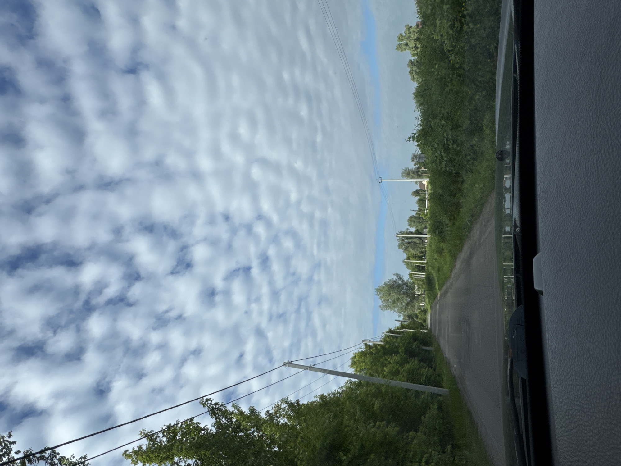 This is a point-of-view shot from inside a vehicle, looking out at a rural road lined with trees and vegetation. The road is paved and curves slightly to the left in the distance. Overhead, a sky filled with white, puffy clouds against a blue backdrop. Telephone poles with wires are visible along the side of the road, extending into the distance. The foreground of the image shows the dashboard of the vehicle, with a windshield wiper visible. The scene suggests a drive through a countryside or semi-rural area, possibly during daytime with overcast conditions.