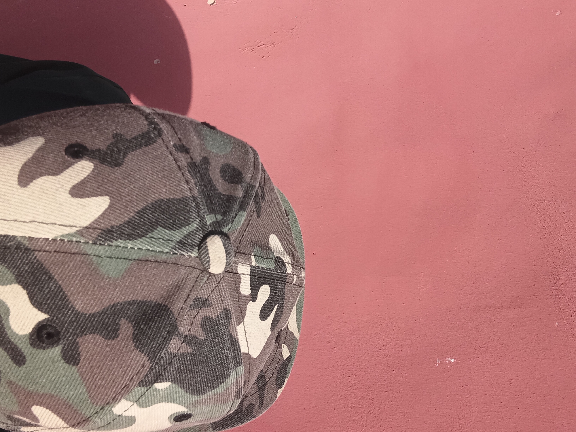 A close-up shot frames the top of a camouflage baseball cap, worn by an unseen individual against a pink, textured wall. The cap, featuring a pattern of greens, browns, and off-white, casts a shadow to the left. The wall exhibits minor imperfections and a slight roughness. The scene appears to be outdoors in Jalingo, Nigeria.