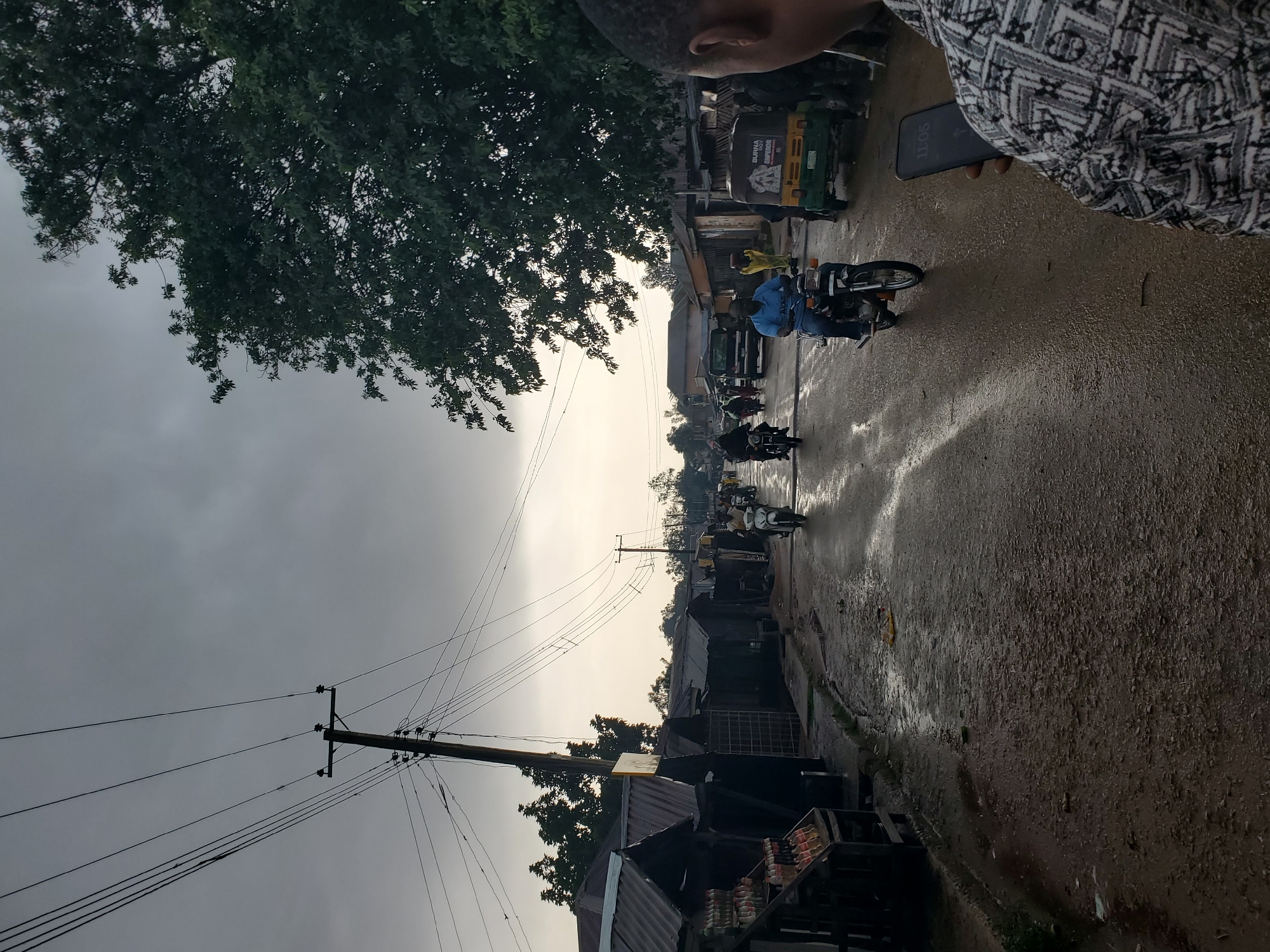 The media file depicts a bustling street scene in Barikin Gidar Kanat, Nigeria, likely during the late afternoon or early evening. The sky is overcast, suggesting the possibility of rain or recent rainfall, as the wet asphalt road reflects the muted light.
The scene is characterized by a lively flow of activity centered around motorcycles, the primary mode of transportation. Several individuals are seen riding motorcycles, navigating the wet street lined with modest buildings and market stalls. In the foreground, a person is holding a smartphone, its screen displaying the time "11:05". Further down the street, a green and yellow trishaw or "Keke Napep" is visible, indicating commercial activity.
The buildings appear to be simple, constructed with corrugated iron roofs and brick or concrete walls, typical of many market areas in the region. The presence of trees on the left side of the image provides some natural element to the urban landscape. Overhead power lines crisscross the sky, supported by wooden poles, a common sight in many communities.
The overall atmosphere suggests a vibrant, everyday moment in a Nigerian town, where commerce and daily life continue despite the weather. The image captures the essence of local transportation and the lively energy of the community.