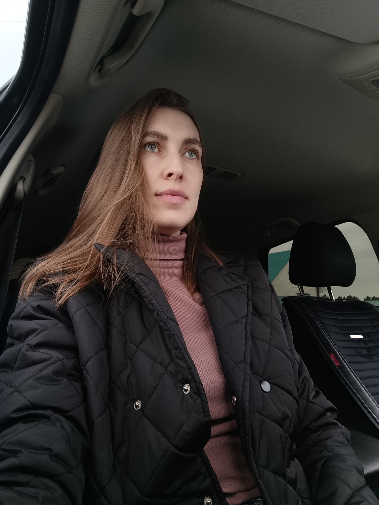 A woman is sitting in the driver's seat of a car. She is wearing a black quilted jacket and a pink turtleneck sweater. She has long brown hair and blue eyes. She is looking up and to the right. The interior of the car is black. The seats are covered in black velvet. There is a window in the background. Outside the window, there is a building with a blue roof, and some trees. The woman's hair is on her left shoulder. The location is likely in the city of Фоки, Russia, based on the prompt.