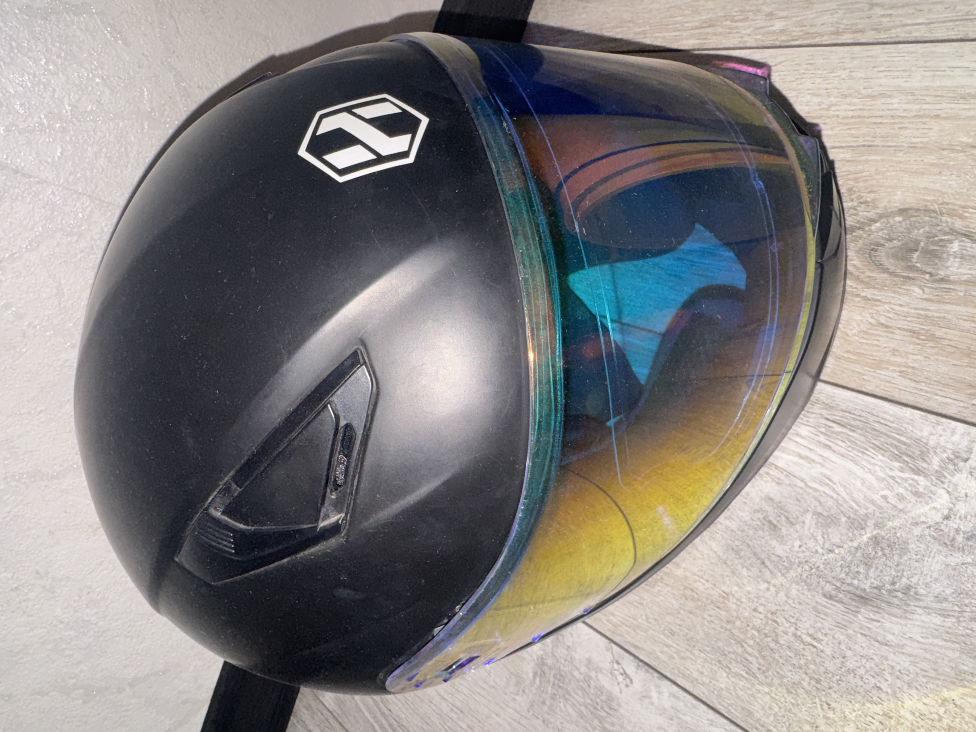 A black motorcycle helmet with a multi-colored visor rests on a light-colored wooden floor against a textured, light-colored wall. The helmet has a white hexagonal logo with a stylized "X" on the right side. A black strap is visible behind the helmet. The visor reflects the surroundings with shades of yellow, blue, and purple. The lighting suggests an indoor setting. There are no people present in the image. The location is not determinable from the image.
