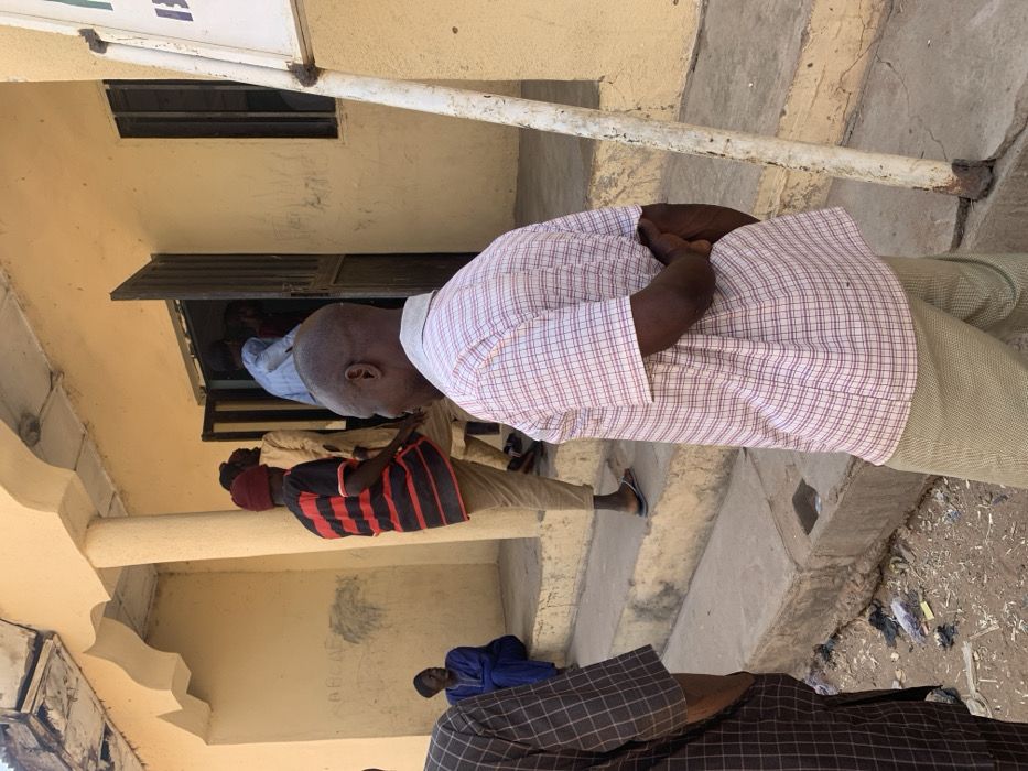 The image captures an outdoor scene in Bojude, Nigeria, depicting multiple individuals near the entrance of a single-story building.
In the immediate foreground, a dark-skinned man with a shaved head stands with his back partially turned towards the camera, angled slightly left. He wears a short-sleeved plaid shirt, primarily white with red and blue checks, and light-colored trousers. His hands are clasped behind his back. To his left, the torso of another man in a dark, checkered long-sleeved garment is visible.
Central to the image, a man wearing a red beanie, a horizontally striped red and black short-sleeved shirt, and light brown trousers with flip-flops, is positioned on a set of three concrete steps leading up to the building's entrance. He faces away from the camera, appearing to ascend or descend the steps. To the far left, another man in a dark blue traditional robe is seated on a lower step, facing right.
The building's exterior is light yellow or tan, showing signs of wear. It features an open doorway with dark metal double doors, through which several indistinct figures are visible, some appearing to enter or exit. One figure inside the doorway wears a light blue shirt, another a light brown traditional outfit. The building has an overhang supported by a rounded, light-colored pillar. Faint, stylized letters "ABCDEF" are visible etched on the wall beneath the overhang to the left of the entrance. A weathered white metal pole, possibly a signpost, stands vertically on the far right.
The ground surface in the foreground is unpaved dirt or sand, littered with various debris including plastic bags and papers. The scene is illuminated by what appears to be bright daylight.