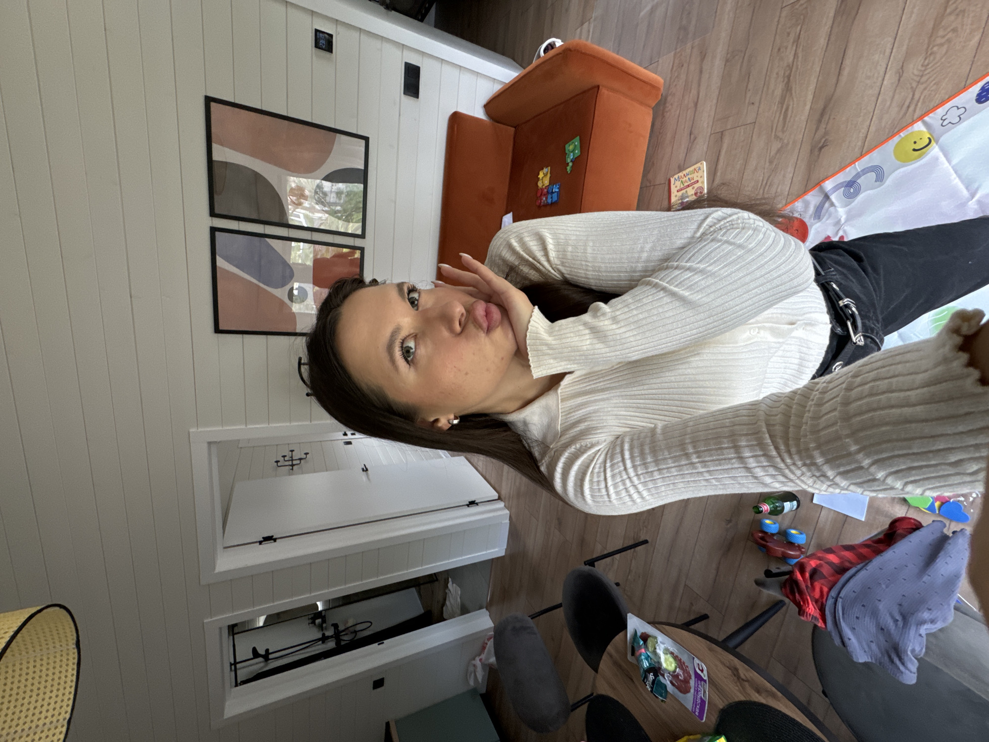 A woman in a white sweater and black pants is taking a selfie in a room. There's an orange chair, wall art, a baby play mat, and a dining table with chairs visible. Toys and clothing are scattered near the table. Based on the location data, the room is located in Литвиново, Russia.