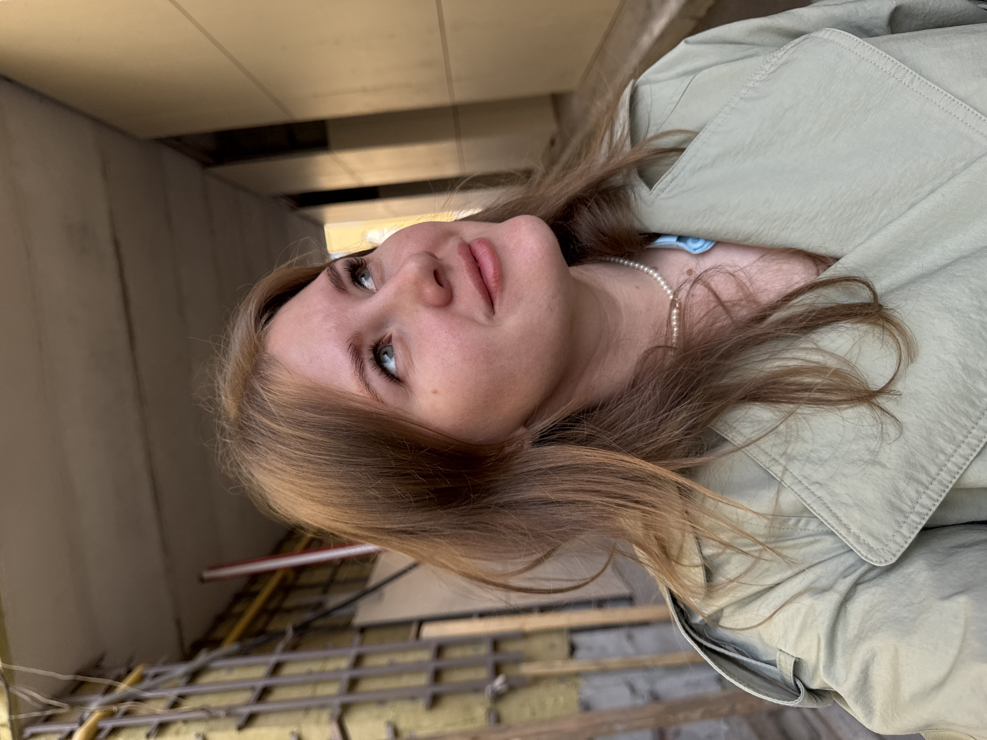 A young, fair-skinned woman with light brown hair and blue eyes poses, looking upwards and slightly to the left. She is wearing a light green trench coat and a pearl necklace. The background suggests an urban environment, possibly a construction site or renovation project, with exposed wooden beams and infrastructure. The photo was taken in the city of Perm, Russia.