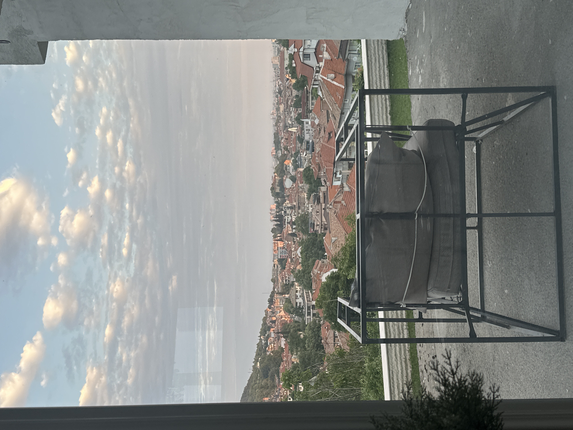 The image presents a panoramic view of the city of Asenovgrad, Bulgaria, observed from an elevated indoor or semi-enclosed vantage point through a glass pane, exhibiting reflections. The time of day appears to be either early morning or late afternoon, characterized by soft, diffused natural light.
The sky occupies the upper half of the frame, displaying a light blue hue interspersed with numerous white and light grey altocumulus clouds. The urban landscape of Asenovgrad extends across the middle and distant background. It features a dense arrangement of residential and commercial buildings, predominantly characterized by traditional red-tiled roofs, amidst abundant green trees and foliage. A prominent, tree-covered hill rises on the left side of the cityscape, while the distant horizon is hazy.
In the mid-ground, on what appears to be an outdoor patio or balcony, a modern outdoor lounge chair with a dark, square-profiled metal frame is centrally positioned. The chair holds two stacked grey cushions, secured with white ropes or ties. The patio floor is a light grey concrete surface. Behind the chair, a low, textured white stone wall is visible, with a small patch of green grass beyond it. A dark, out-of-focus plant is visible in the lower left corner, indicating an indoor element. No individuals are present in the scene.
