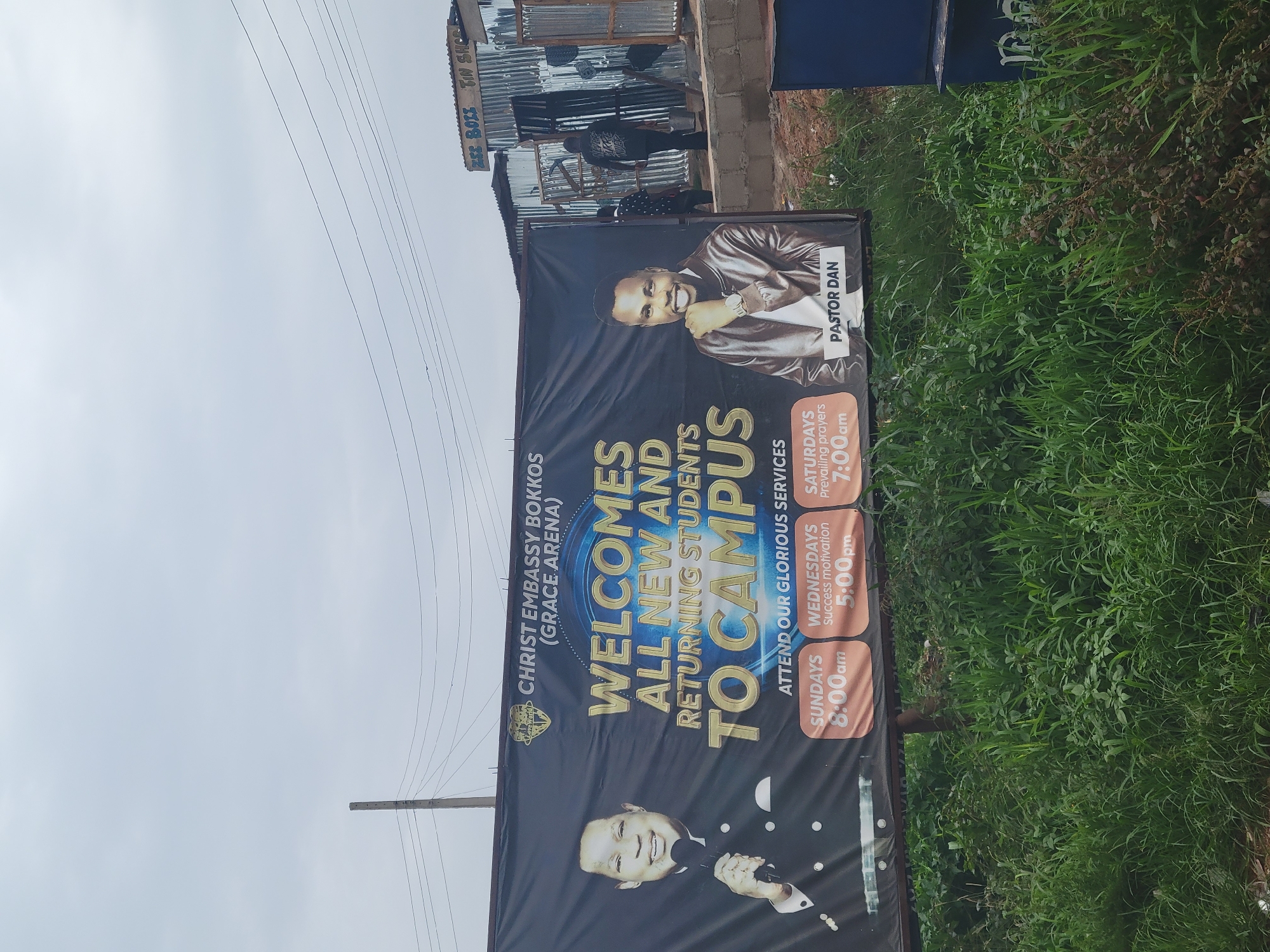 The image shows a large banner with text that reads "CHRIST EMBASSY BOKKOS (GRACE ARENA) WELCOMES ALL NEW AND RETURNING STUDENTS TO CAMPUS". The banner also lists service times: Sundays at 8:00 am, Wednesdays at 5:00 pm (Success motivation), and Saturdays at 7:00 am (Prevailing prayers). Two men are featured on the banner: one labeled "PASTOR DAN" and another smiling man at the bottom.
In the background, a corrugated metal structure is visible, possibly a shop or a building under construction. There are also power lines and lush green vegetation, including tall grass and bushes, to the right of the banner. The sky appears overcast, suggesting a cloudy day. The overall scene appears to be an outdoor setting, likely in a community or campus area, promoting a religious event or organization. The banner's message and imagery suggest a welcoming and encouraging atmosphere. The text "ΖΕΣ BOLI TIN SHO" is also visible on the background structure, though its meaning is unclear in this context.