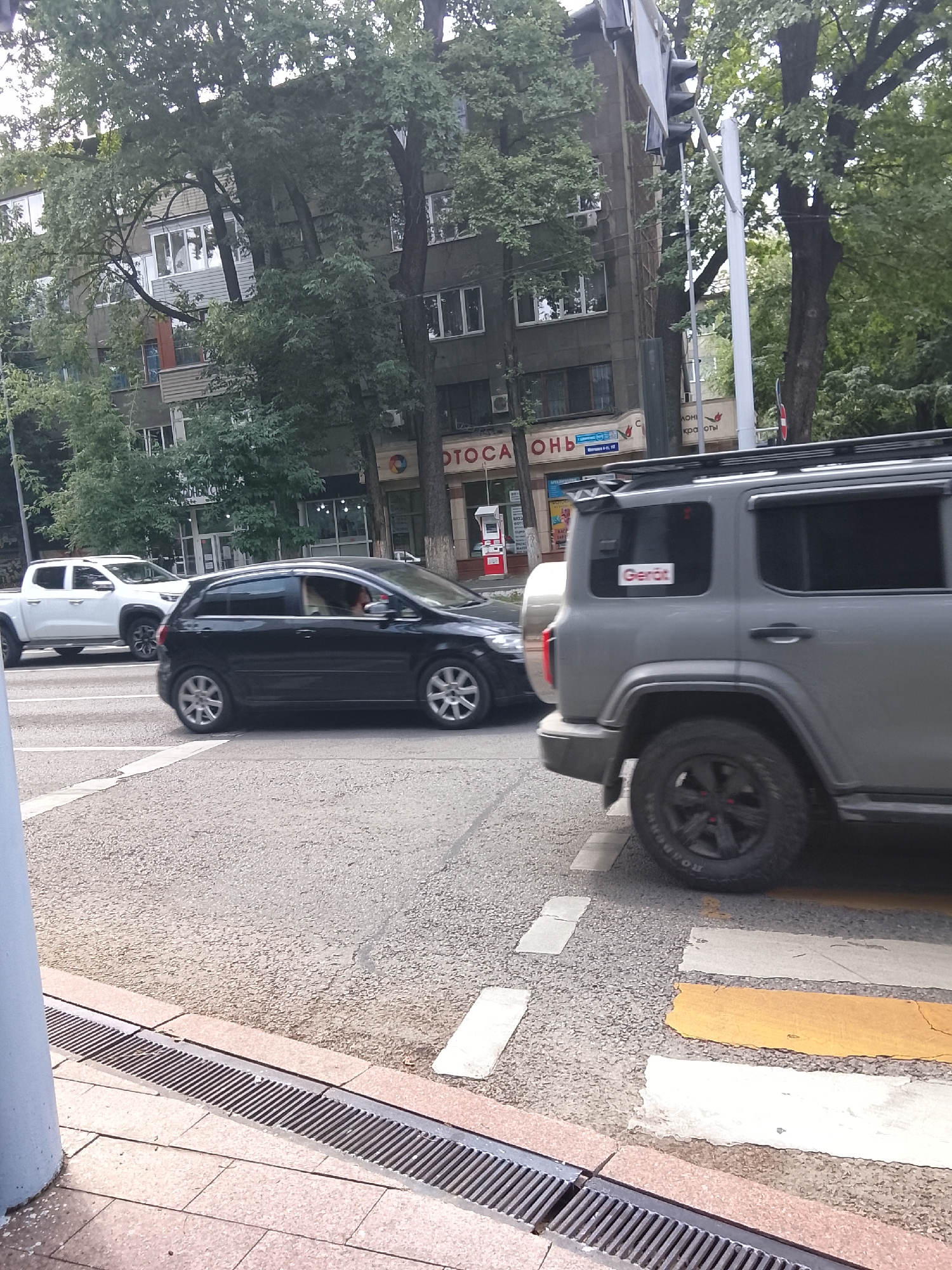 In Almaty, Kazakhstan, on a city street with buildings and trees lining the road, several vehicles are present. A gray SUV is positioned in the foreground, facing right. A black hatchback is to the left of the SUV, also facing right. Further to the left, a white pickup truck is visible, also facing right. A pedestrian crossing with yellow and white stripes is marked on the asphalt. In the background, buildings and trees dominate the scene. The weather appears to be overcast.