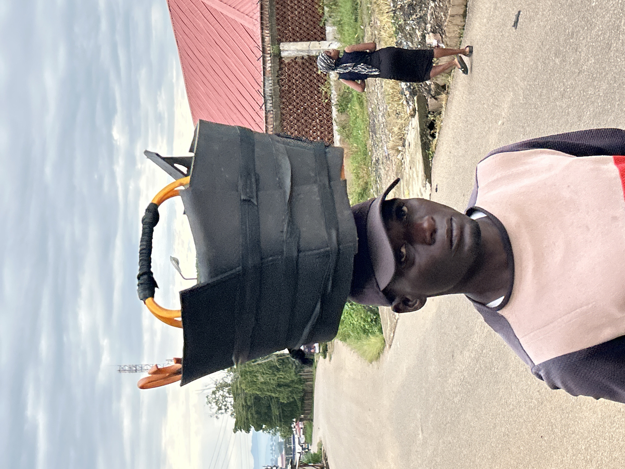 In the city of Jos, Nigeria, a man wearing a dark baseball cap and a long-sleeved shirt with contrasting beige and dark gray panels balances a large, black, rectangular object on his head. The object appears to be a heavy-duty bucket or container, possibly made of tarp or thick plastic, with orange handles that are wrapped in black material. In the background, a woman in a black sleeveless dress and a headscarf walks away from the camera down a paved road. A red-roofed building with a patterned brick wall is visible to the right, and a telecommunications tower is in the distance. The sky is overcast with clouds, and there is vegetation and debris along the side of the road.