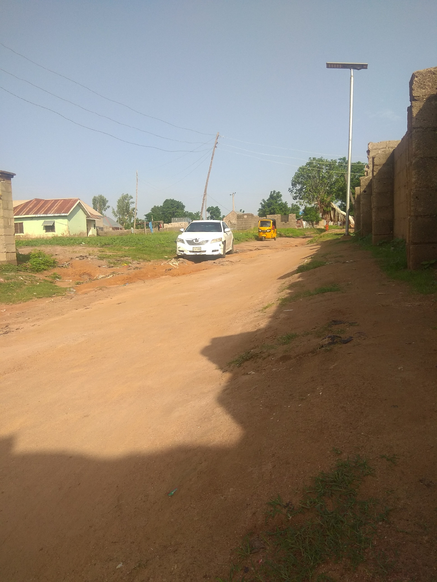 This image depicts a dirt road in Jalingo, Nigeria. A white sedan is parked on the left side of the road, facing away from the camera. Further down the road, a yellow auto-rickshaw is visible. On the left side of the road, there is a light green building with a brown roof. On the right side, a tall concrete wall lines the road, and a solar-powered street lamp stands near it. The sky is clear and blue. The environment appears somewhat unkempt, with scattered debris and vegetation along the roadside.