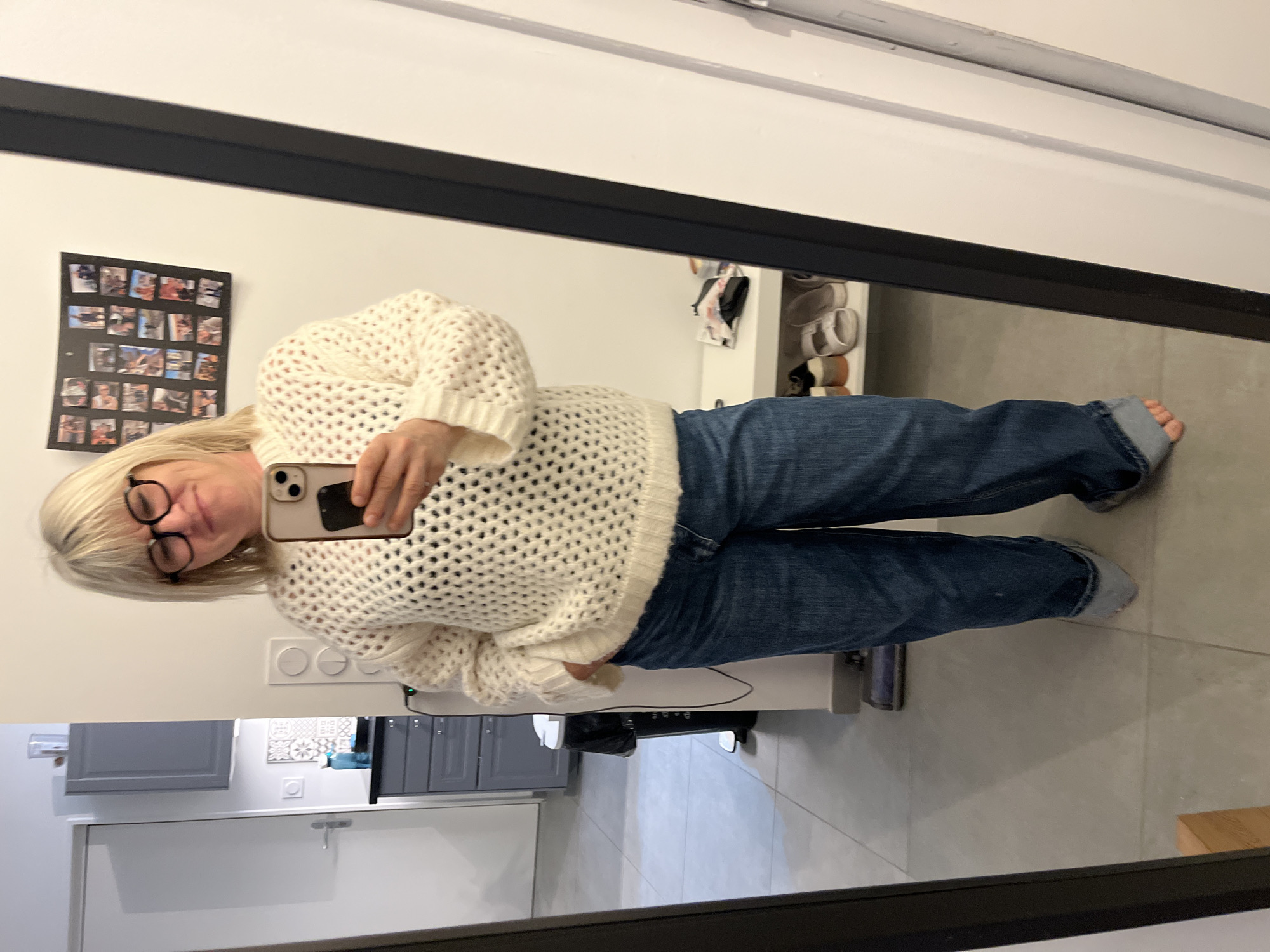 A woman is taking a selfie in a mirror. The mirror is in a room with gray cabinets and drawers. There is a white shelf with shoes on it. The woman has blonde hair and is wearing glasses, a white sweater, and blue jeans. There is a collage of photos on the wall behind her. The image might be in or around Chambœuf, France, but that cannot be determined based on the image alone.