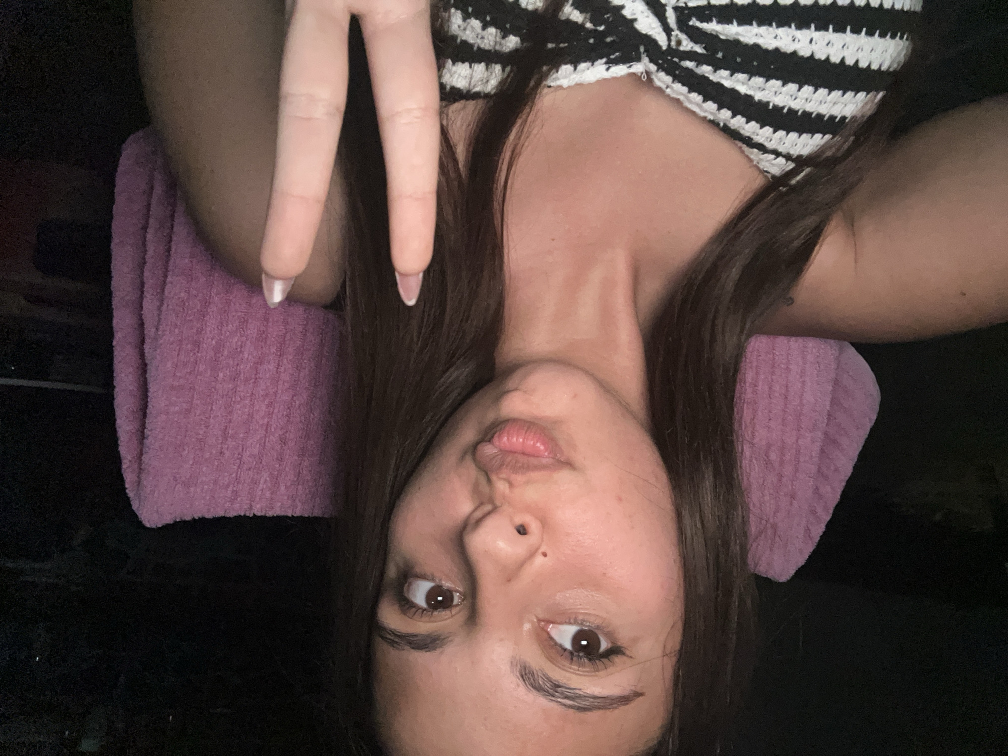 The image features a young adult female with long, dark brown hair and brown eyes, looking slightly upward and to the viewer's right. She has a small mole on the bridge of her nose and her lips are pursed. She is wearing a black and white striped crocheted top. Her right hand (viewer's left) is raised, forming a "peace sign" with her index and middle fingers extended. Her fingernails are almond-shaped with light-colored polish and white tips. Behind her, a pink, fuzzy, ribbed fabric, possibly a cushion or part of a chair, is visible. The background is predominantly dark and indistinct, indicating a dimly lit environment. The lighting appears to be a direct, frontal source. The location is in the city of Unknown, Unknown.