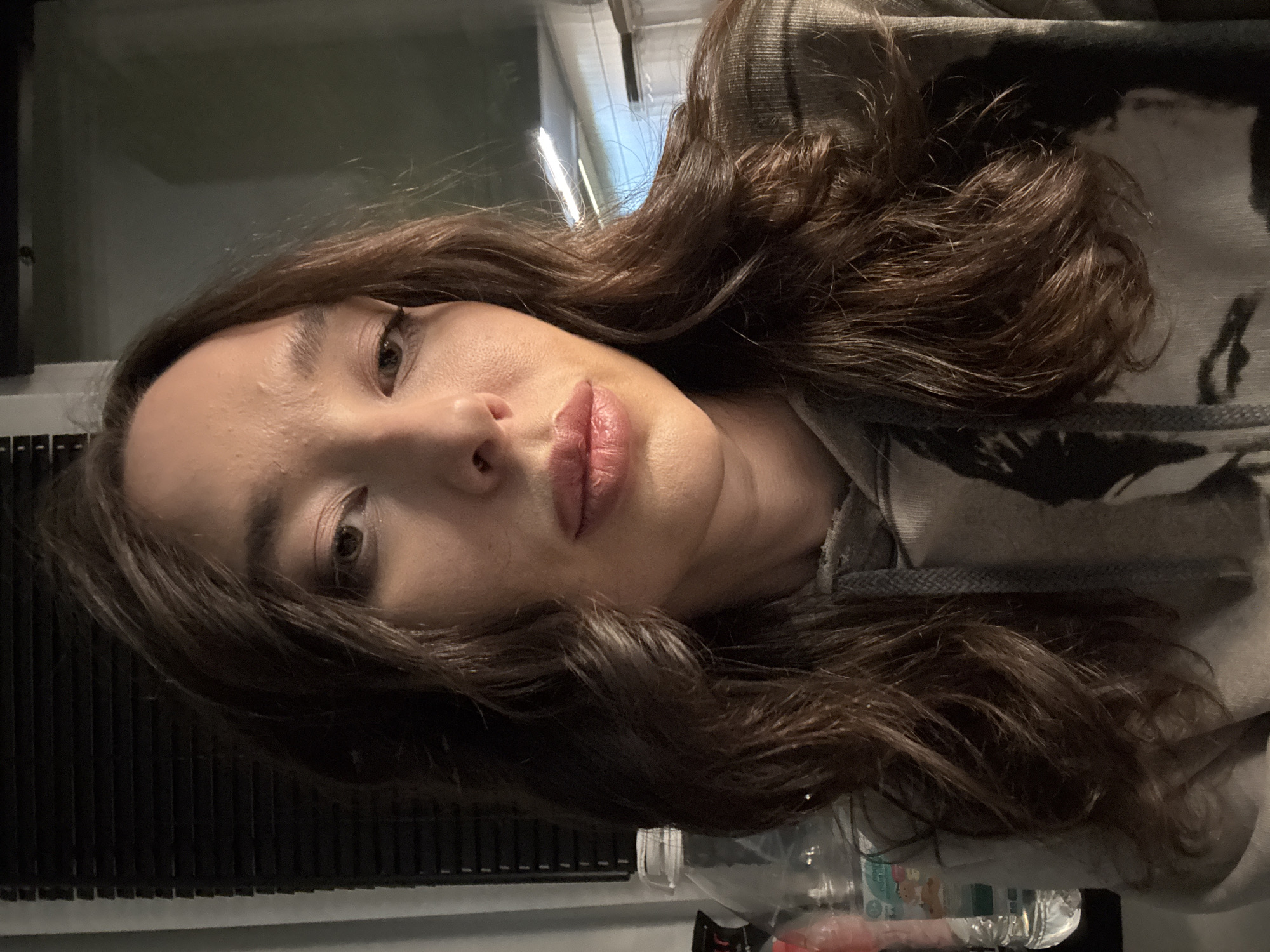 A close-up, angled shot of a person with long, wavy brown hair wearing a hooded sweatshirt. The person has fair skin, light eyes, and pink lips. The background is blurred and indistinct but appears to be an interior space. There is no indication of Moscow, Russia in this image.