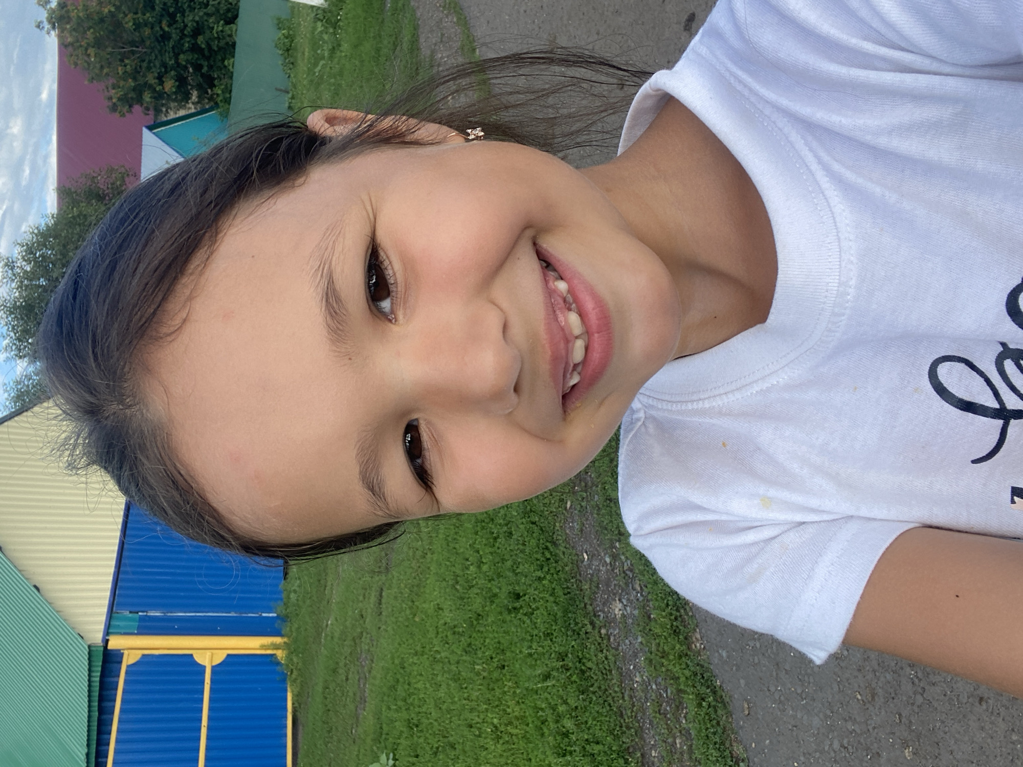 The image is a portrait of a young girl with dark hair, brown eyes, and a gap in her front teeth, smiling. She is wearing a white t-shirt. The background features a blue fence with yellow accents, green grass, and houses with colorful roofs. The scene appears to be in a rural or suburban setting, possibly the city of Maloarslangulovo, Russia, as suggested by the architectural style and vegetation. The lighting suggests it is daytime, with natural light illuminating the girl's face.